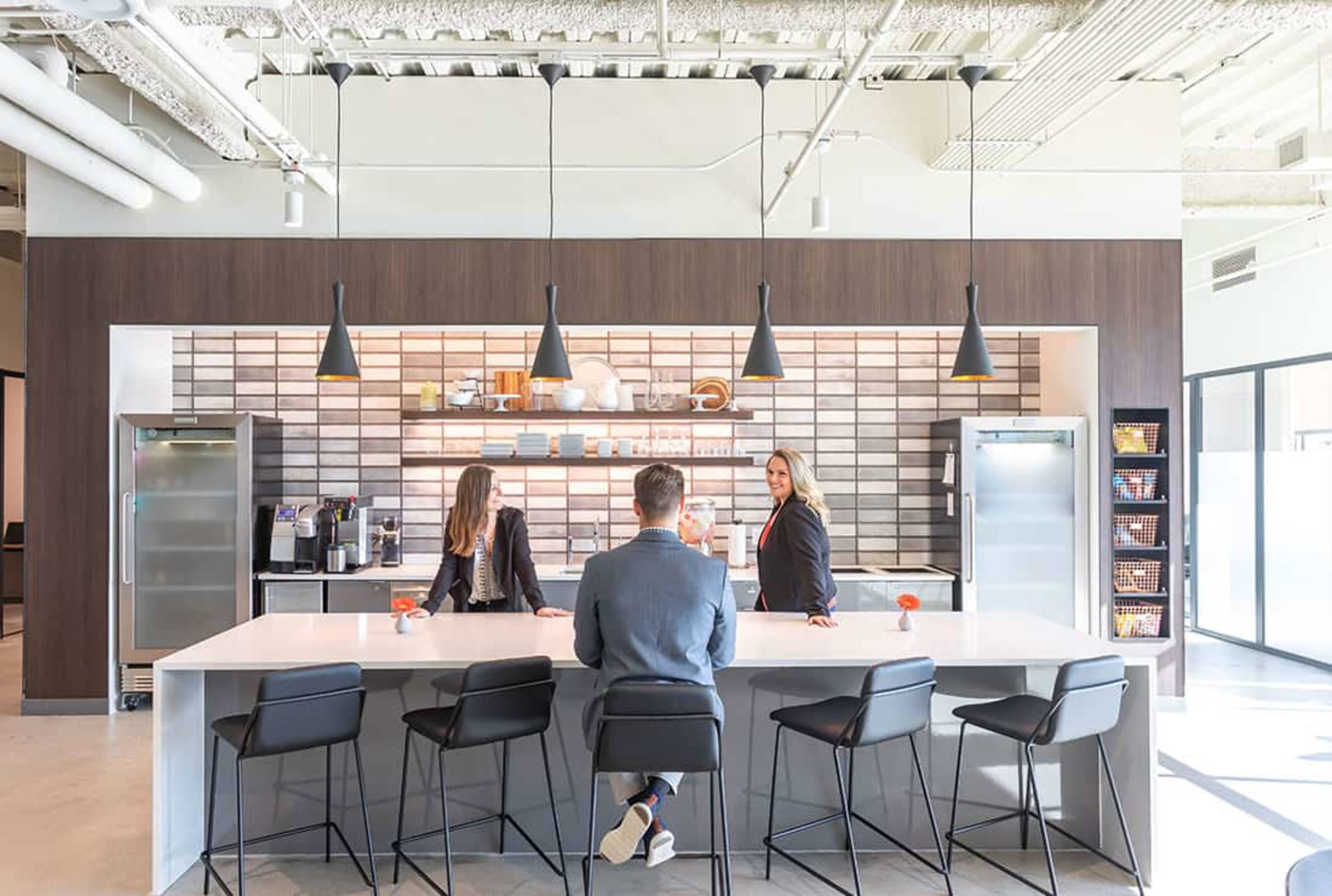Two women and a man converse at a modern kitchen island in an office setting, surrounded by sleek cabinetry and appliances.