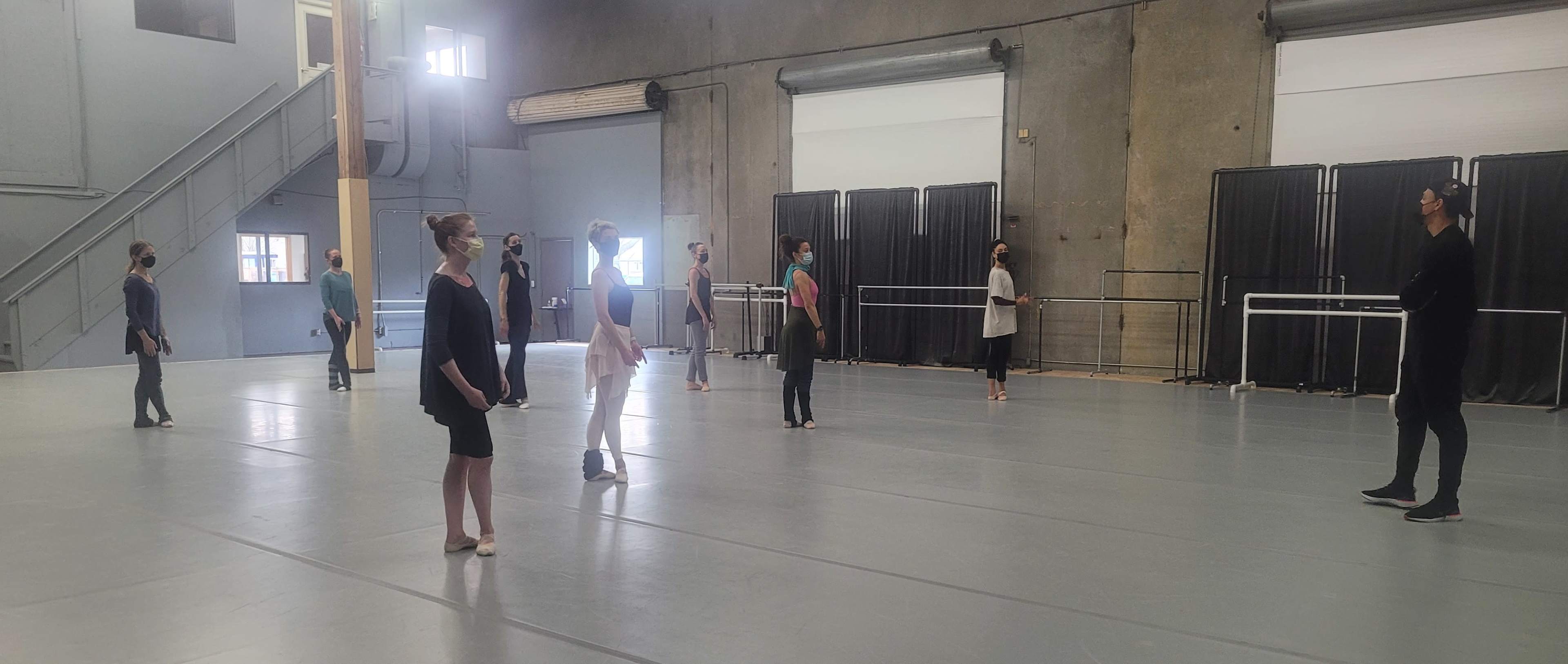 A group of dancers in practice attire stands spaced apart in a large dance studio with mirrors and ballet barres.