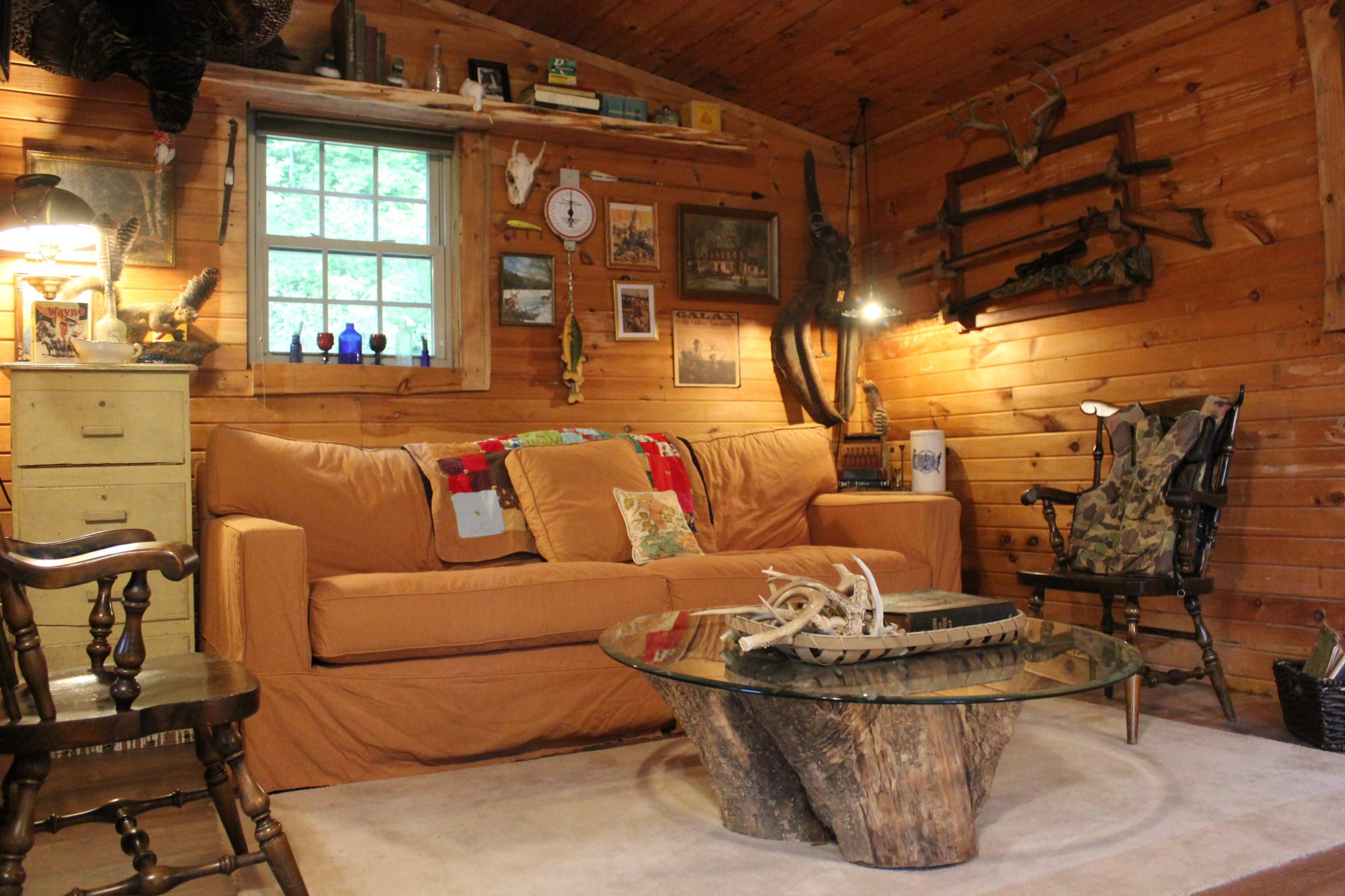 The image shows a cozy wooden cabin interior with a beige couch, a glass coffee table made from a tree stump, and rustic decor on the walls.