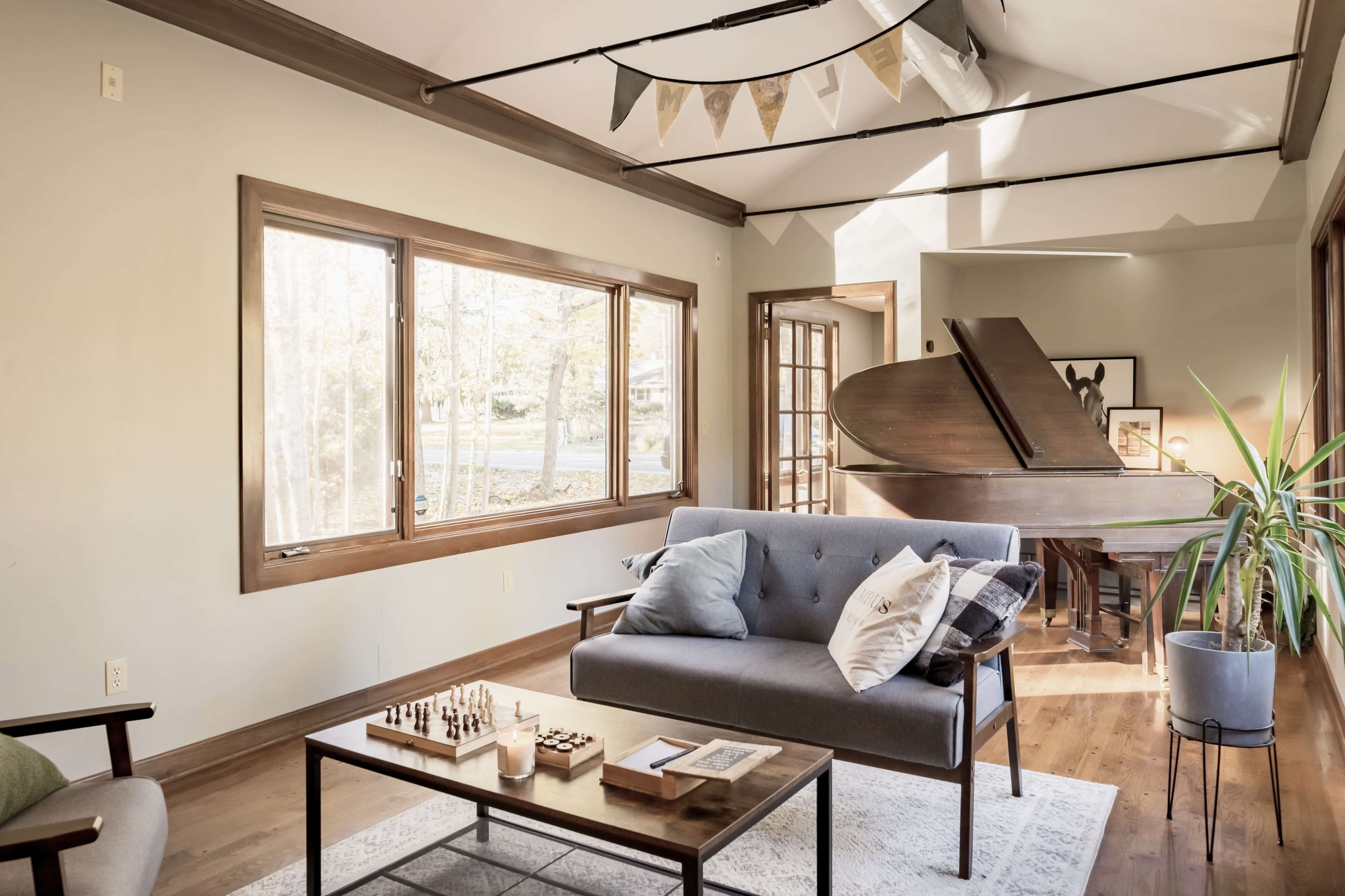 A cozy living room features a gray sofa, a wooden coffee table with a chess set, and a grand piano near large windows that let in natural light.