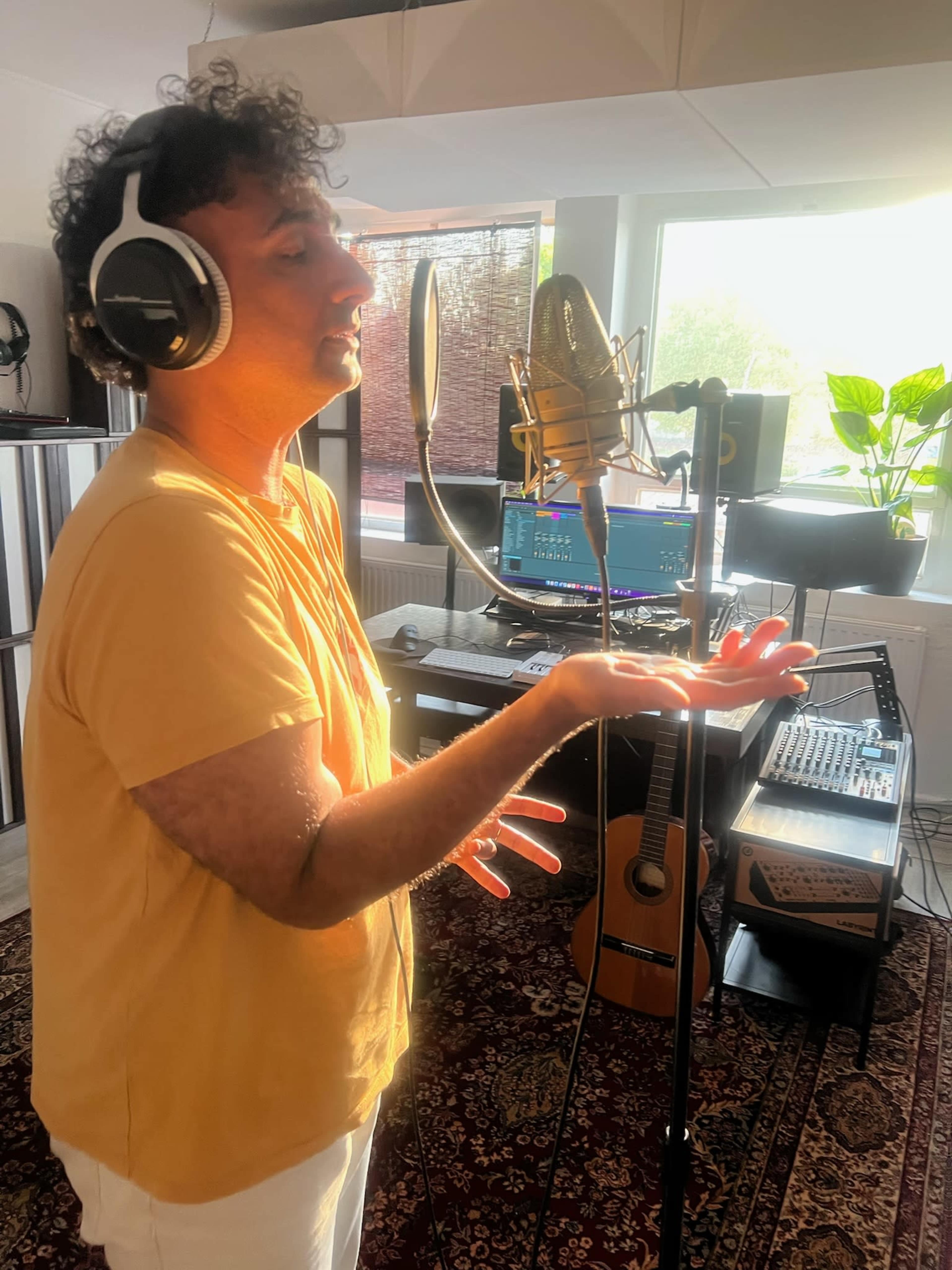 A person is singing into a microphone in a recording studio, with a computer and musical equipment visible in the background.