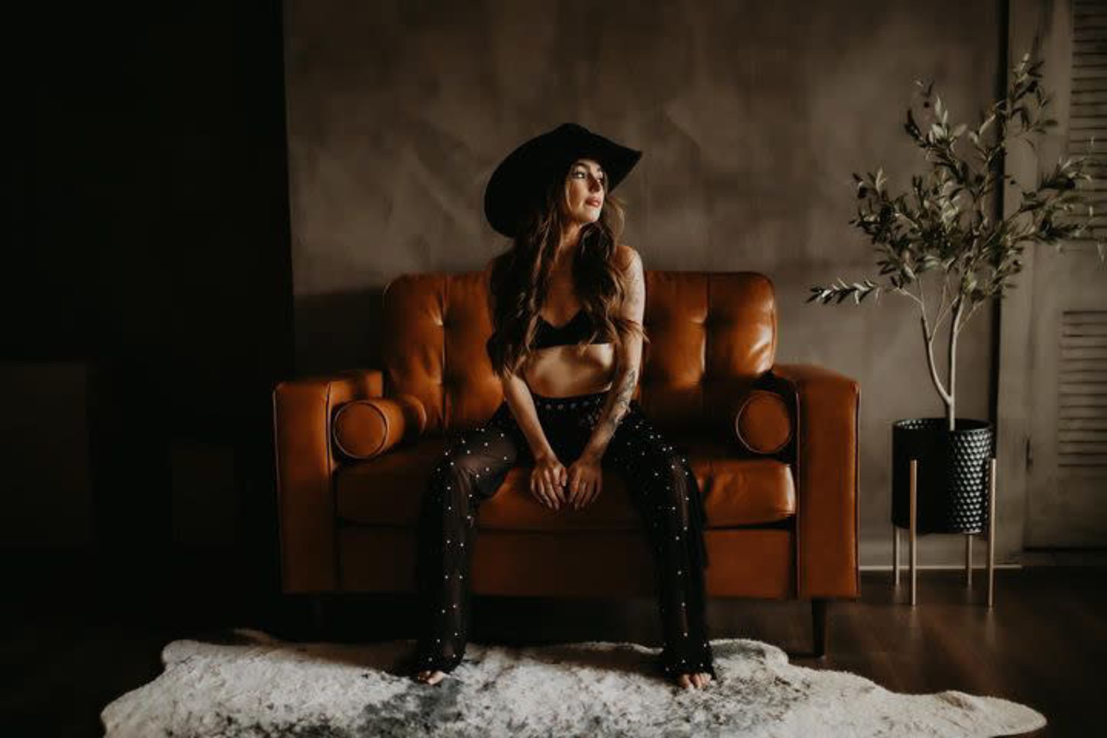 A woman wearing a black bra and wide-brimmed hat sits on a brown leather couch with an olive branch plant nearby.