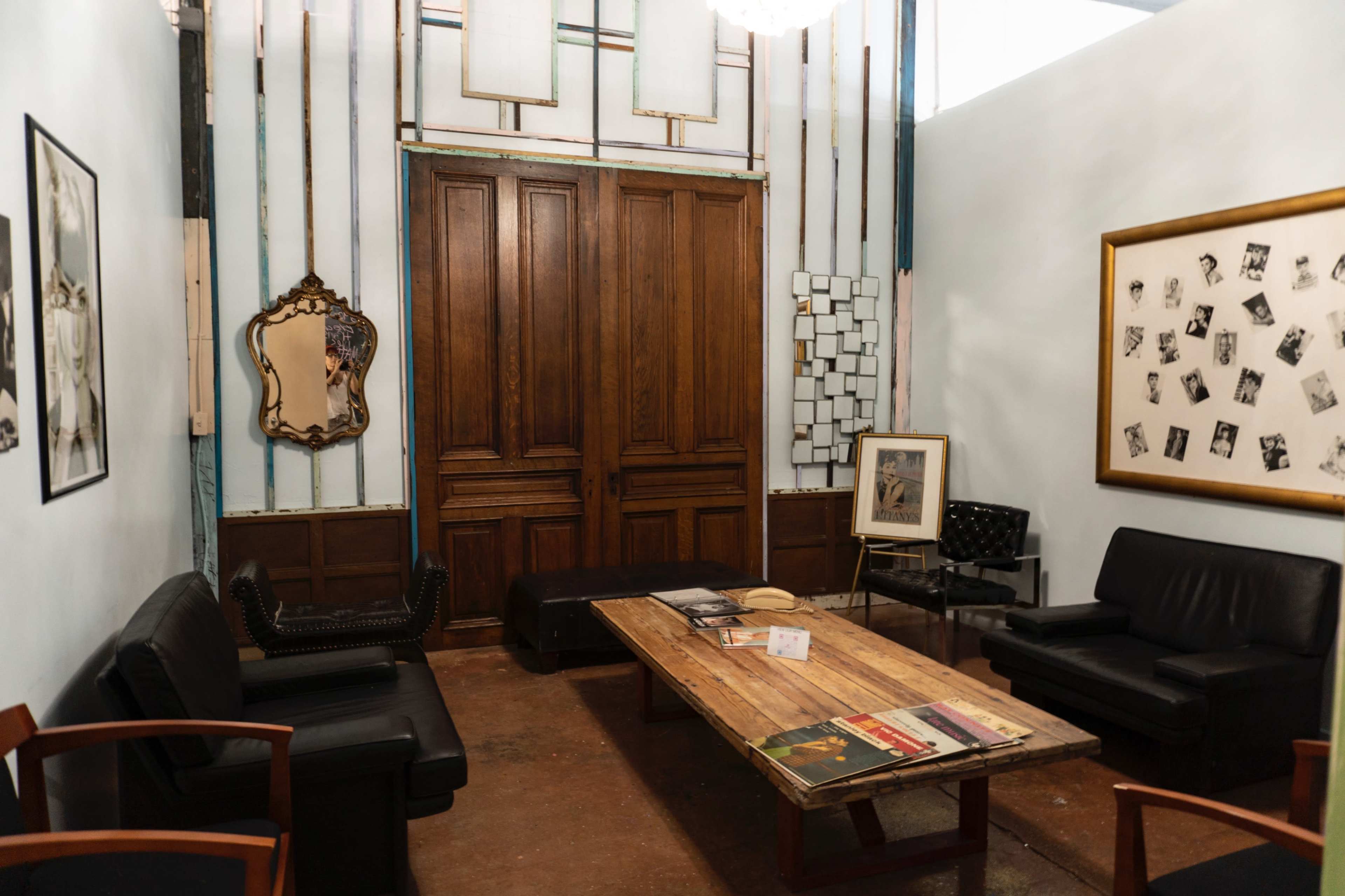 The image depicts a cozy waiting room with wooden doors, two black sofas, a wooden coffee table, and various framed artworks on the walls.