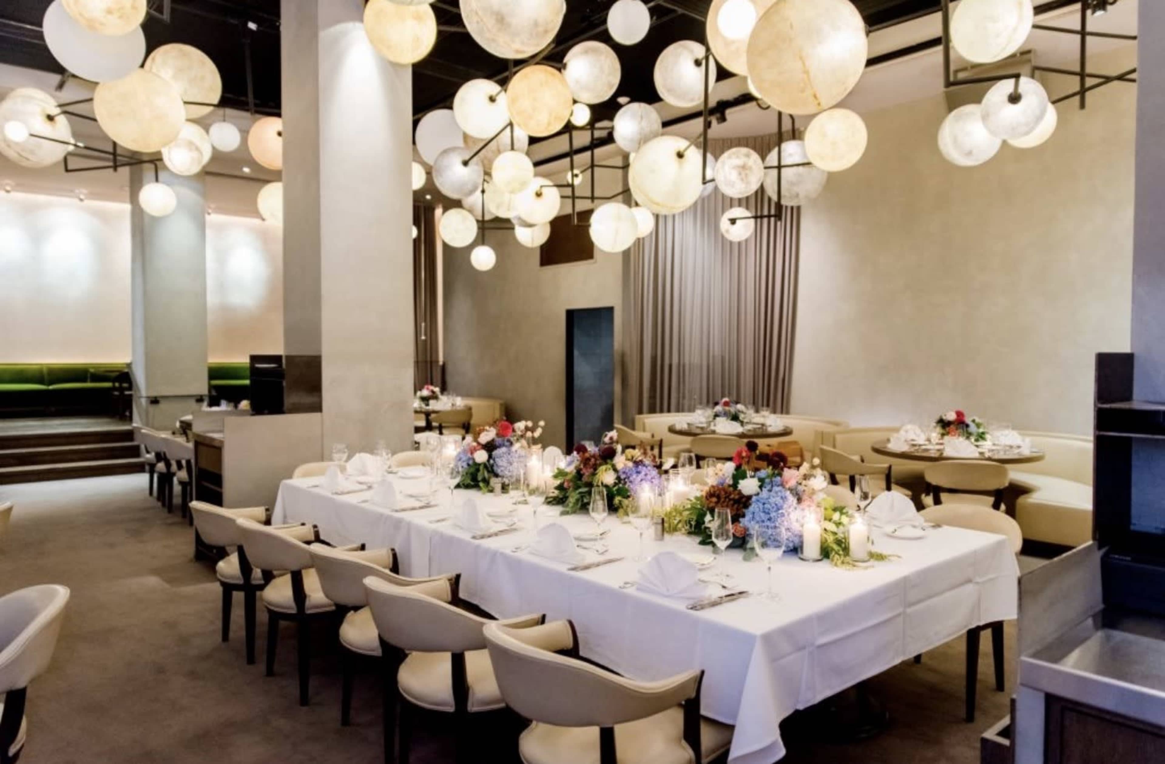 A long dining table is set in a modern restaurant, illuminated by round glass pendant lights and adorned with floral centerpieces.
