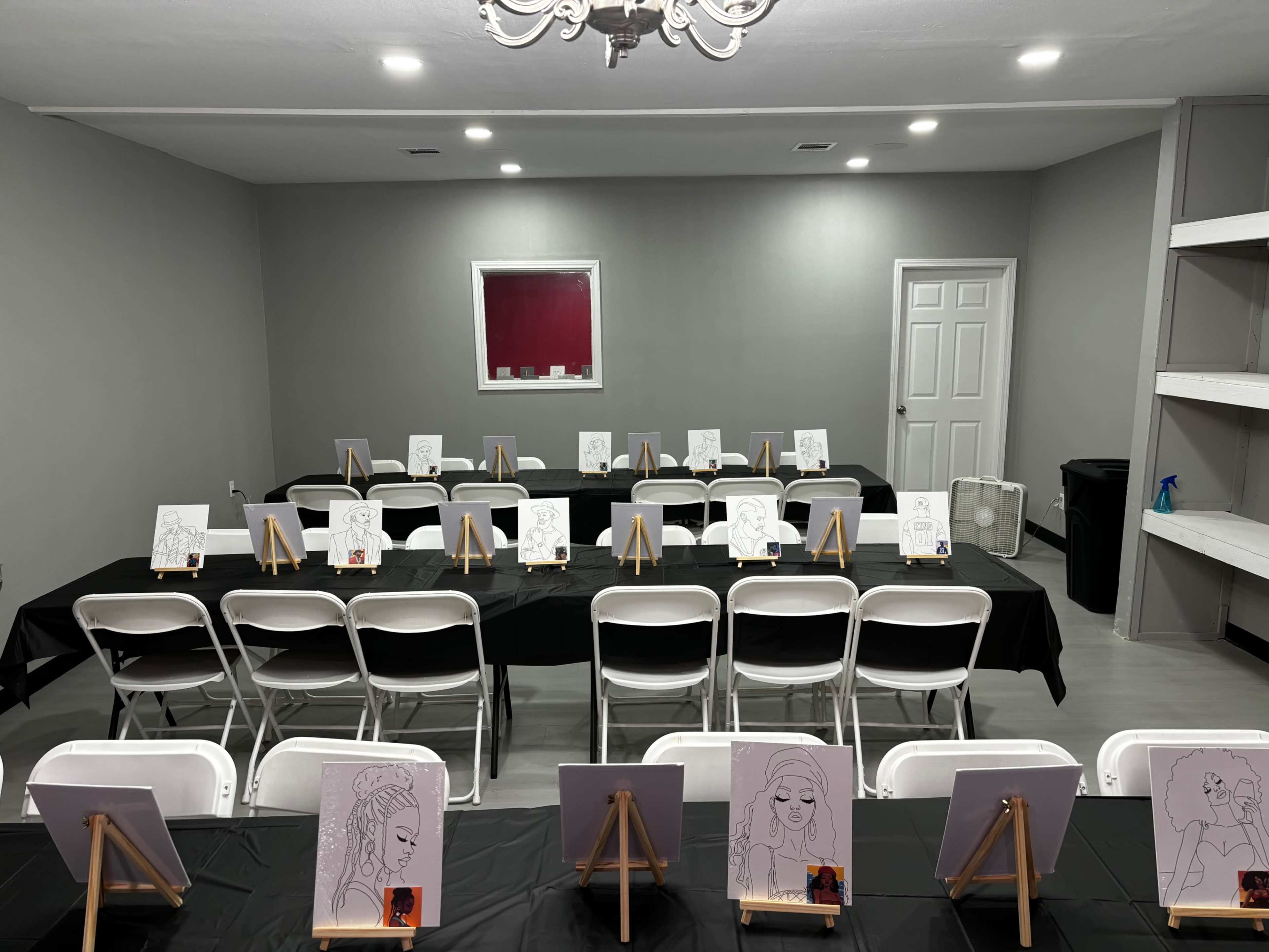 The room features rows of white chairs facing easels with black and white sketches on each, arranged on tables covered with black cloth.
