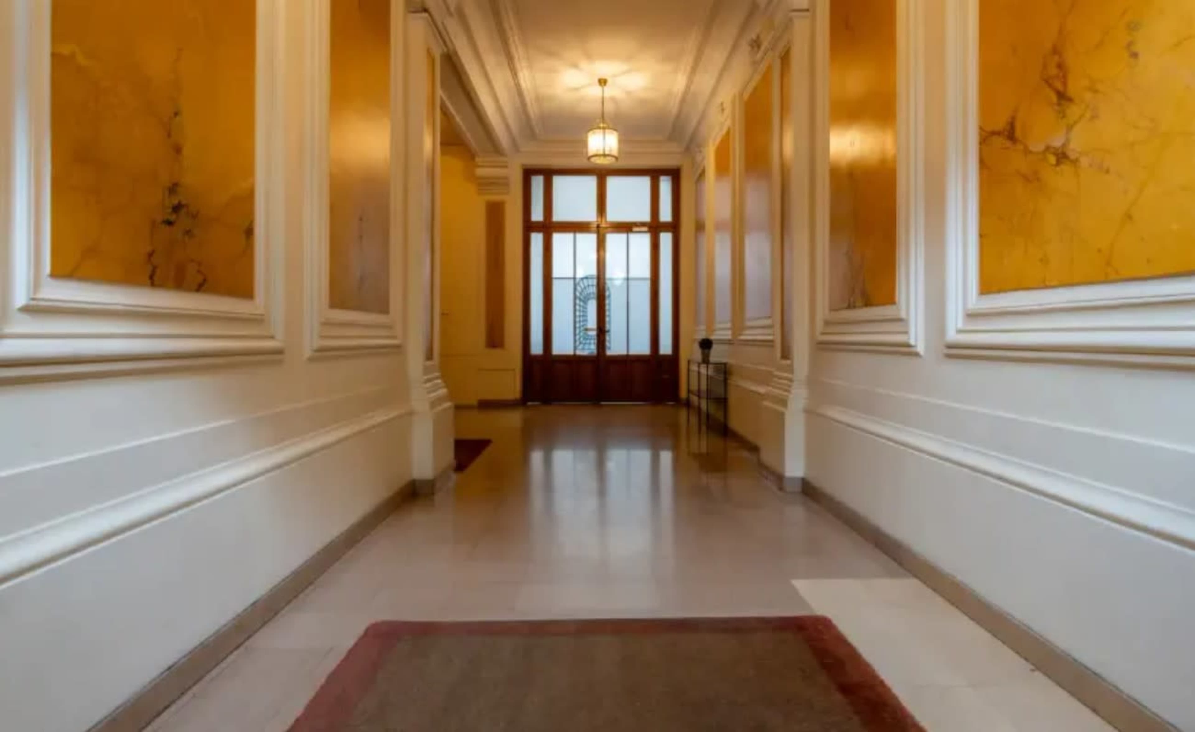 The image shows a long hallway with yellow marble walls, a chandelier, and a glass-divided door at the far end.