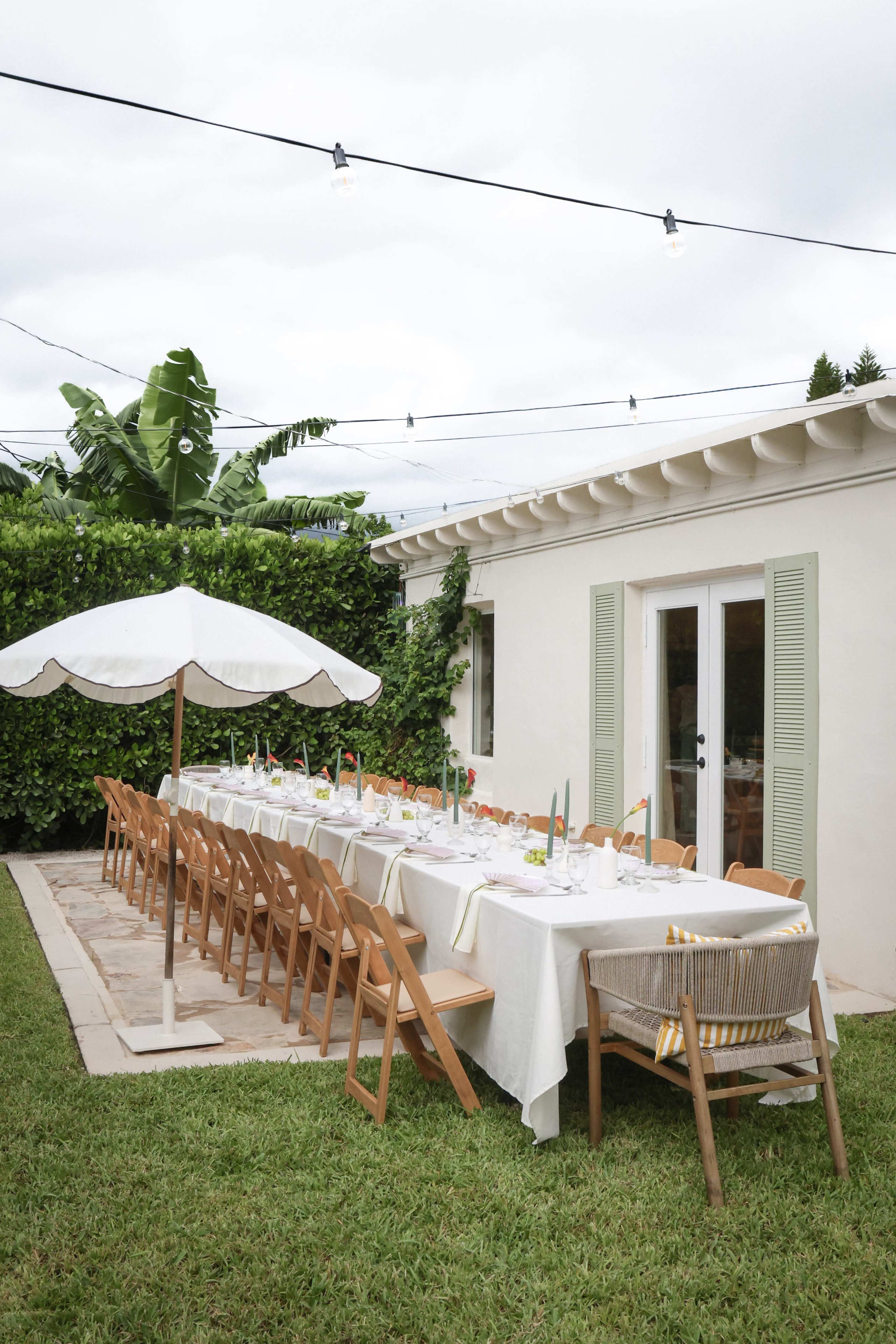 A long dining table is set outside under string lights, with a white umbrella nearby, surrounded by greenery and a modern house.