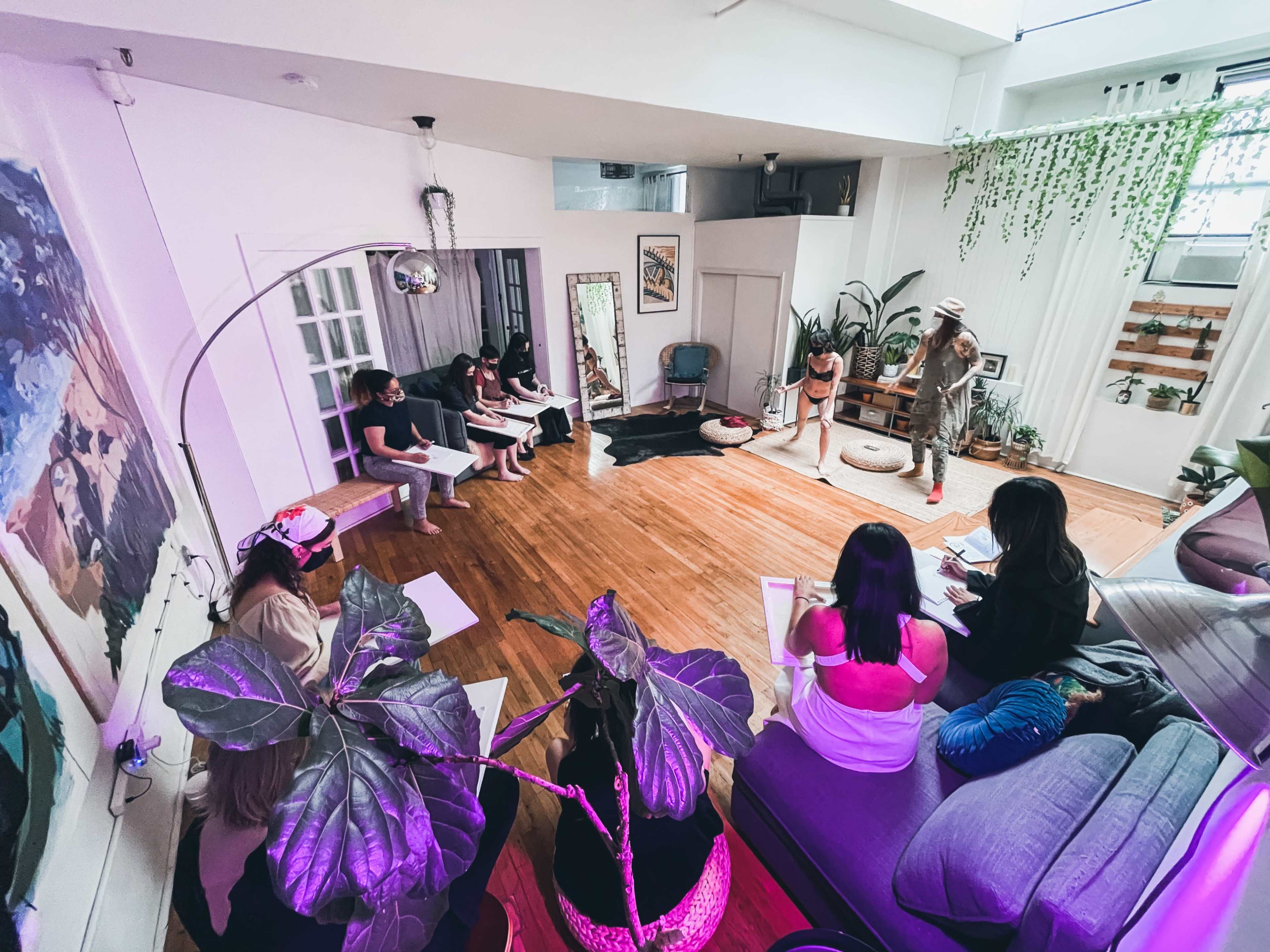 A group of people sits in a semi-circle on the floor of a bright room with plants and art, while one person stands in the center.