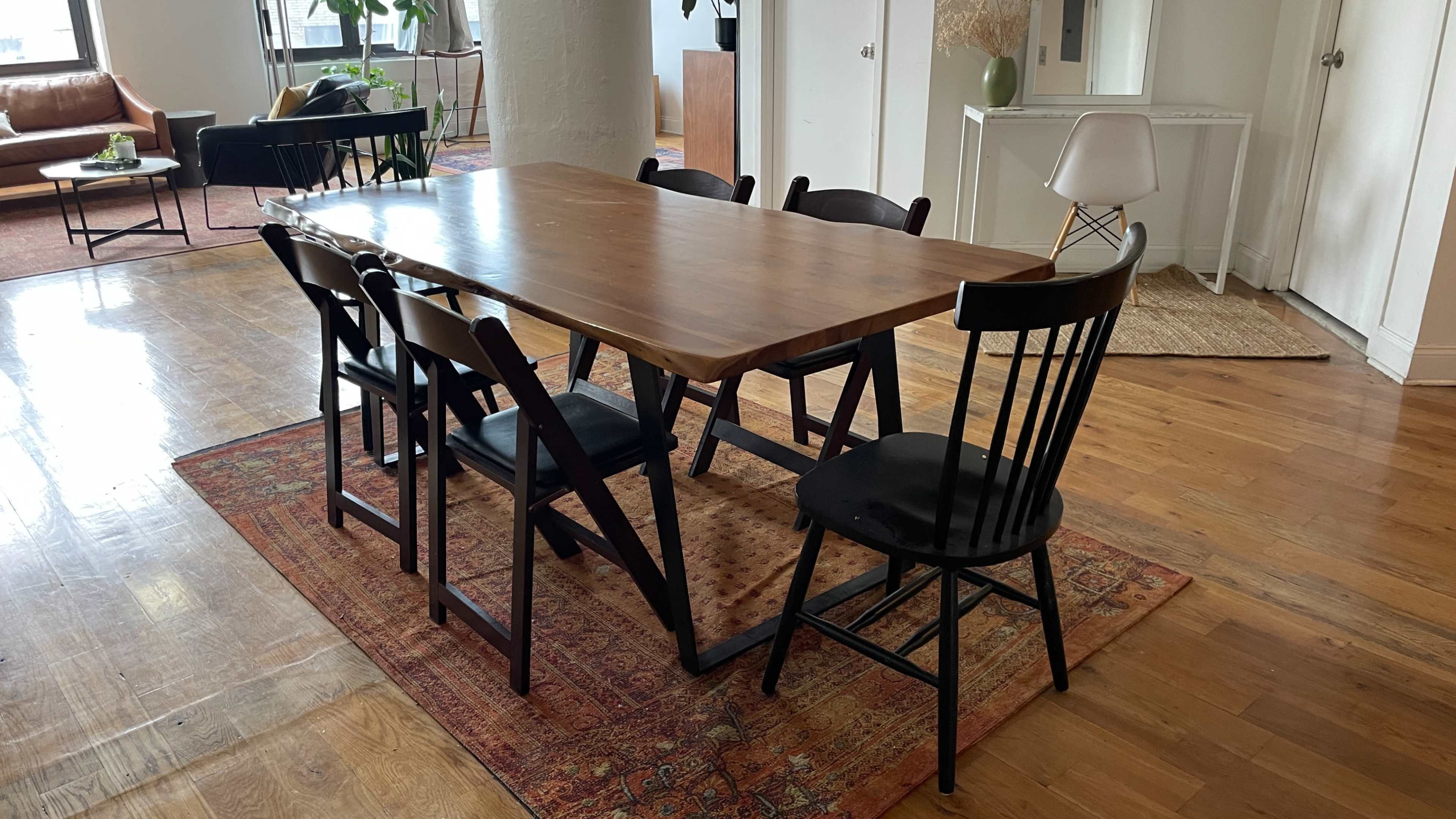 A wooden dining table with black chairs is set on a patterned rug in a well-lit room with hardwood floors.