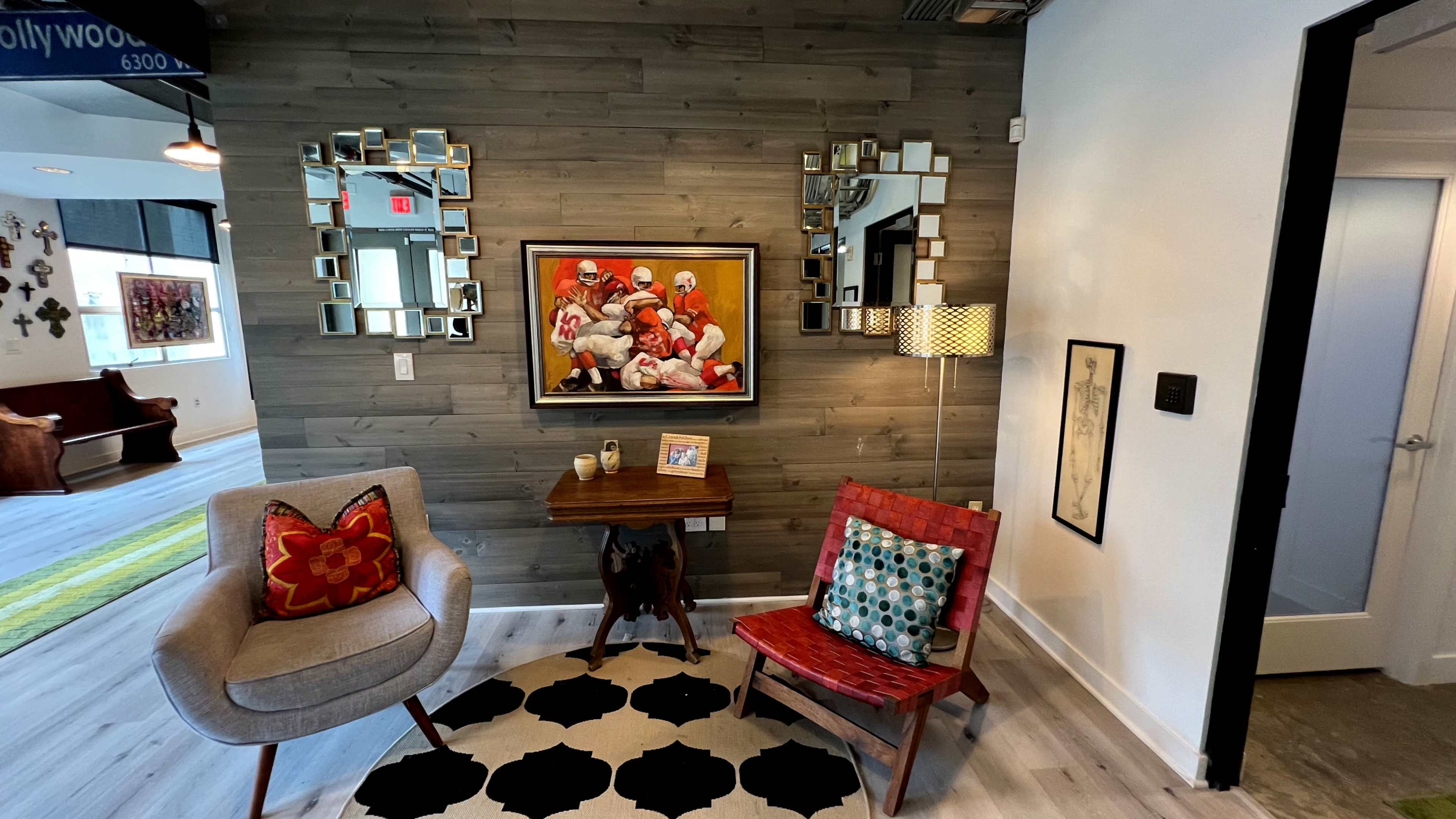 The image shows a modern entryway with wooden walls, two decorative mirrors, a colorful painting, a small table, and two distinct chairs on a patterned rug.