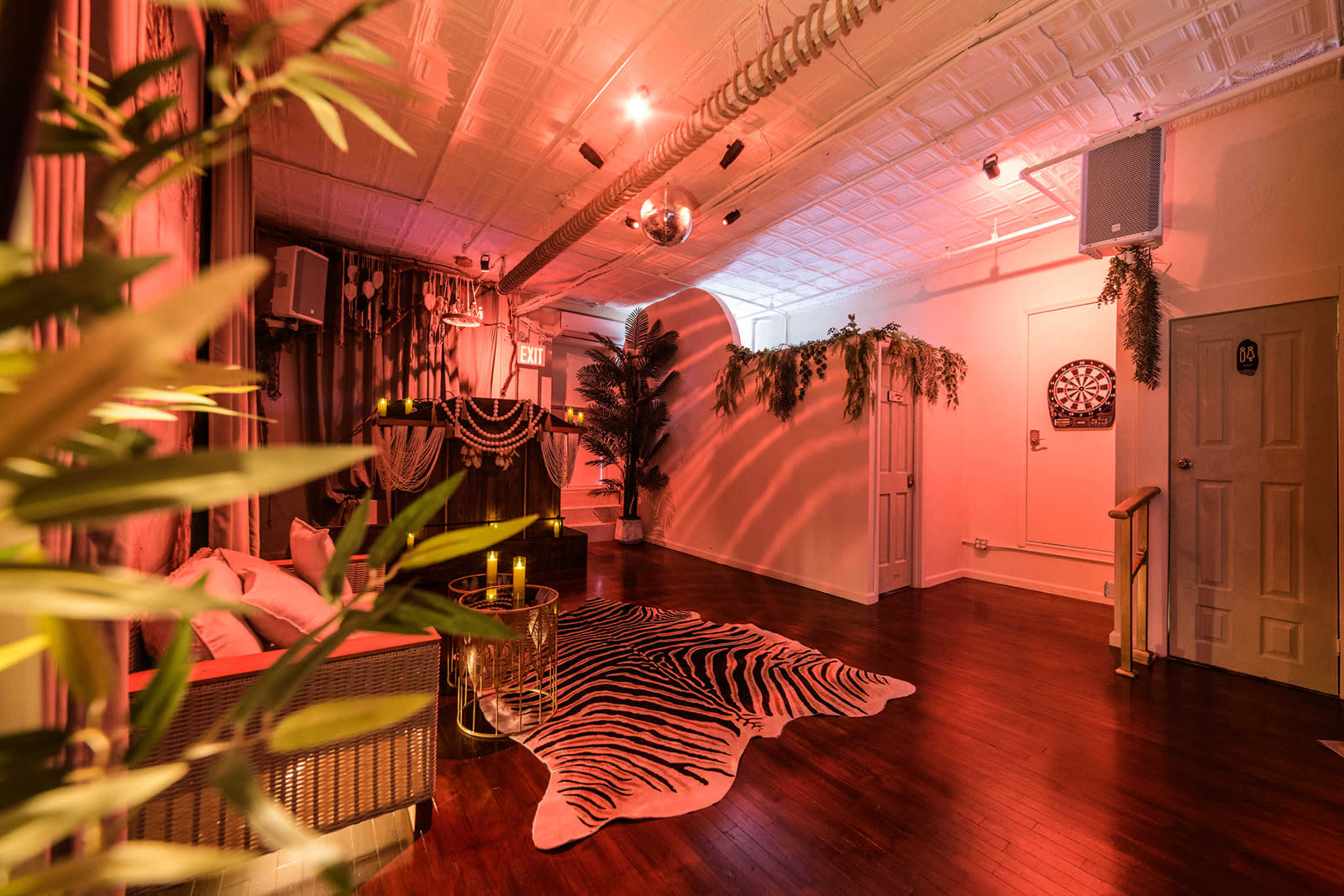 The image shows a stylishly decorated indoor space featuring warm lighting, a zebra-print rug, comfortable seating, and plants against a backdrop of exposed brick and wood finishes.