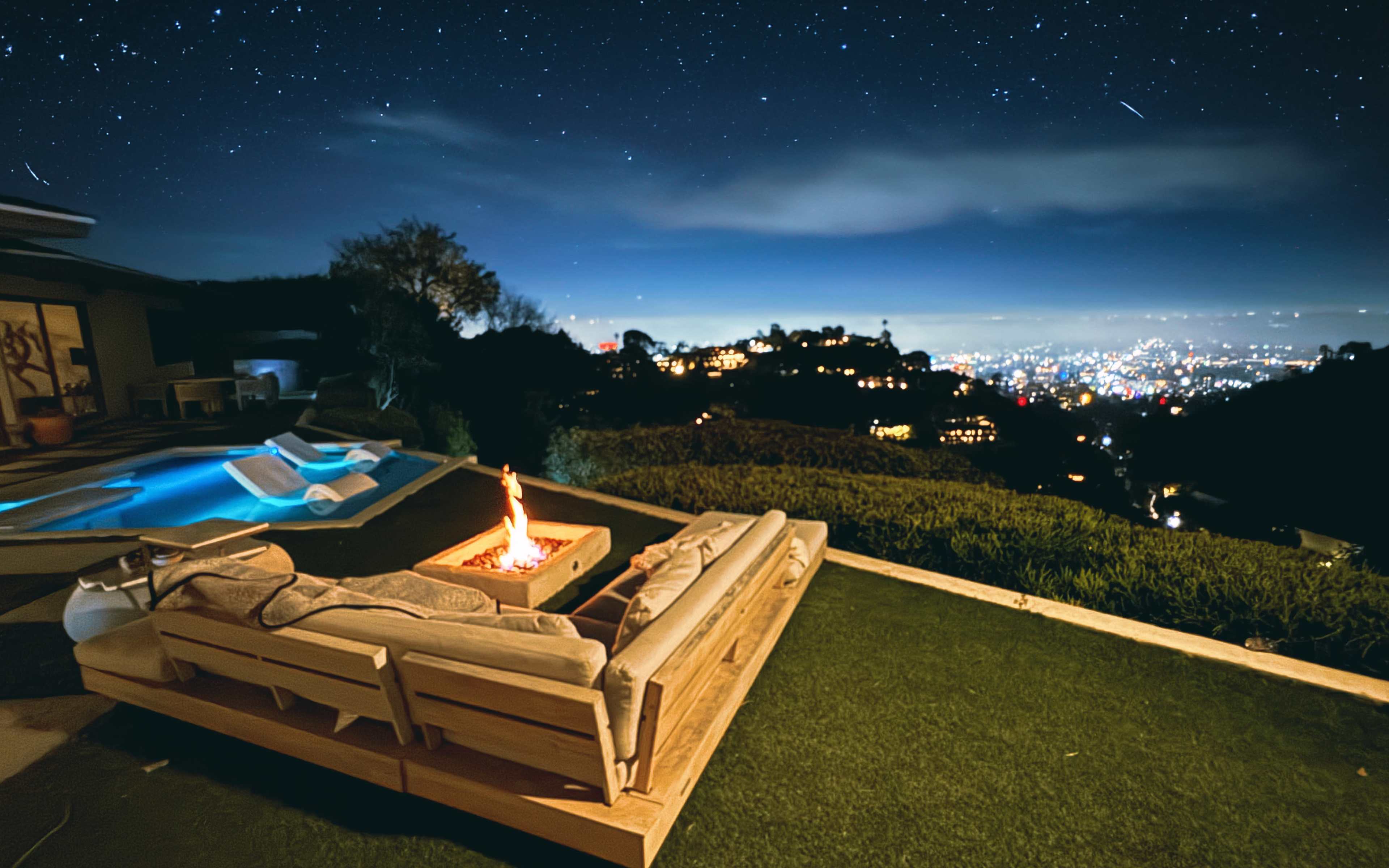 A modern outdoor seating area with a fire pit overlooks a city skyline under a starry night sky.