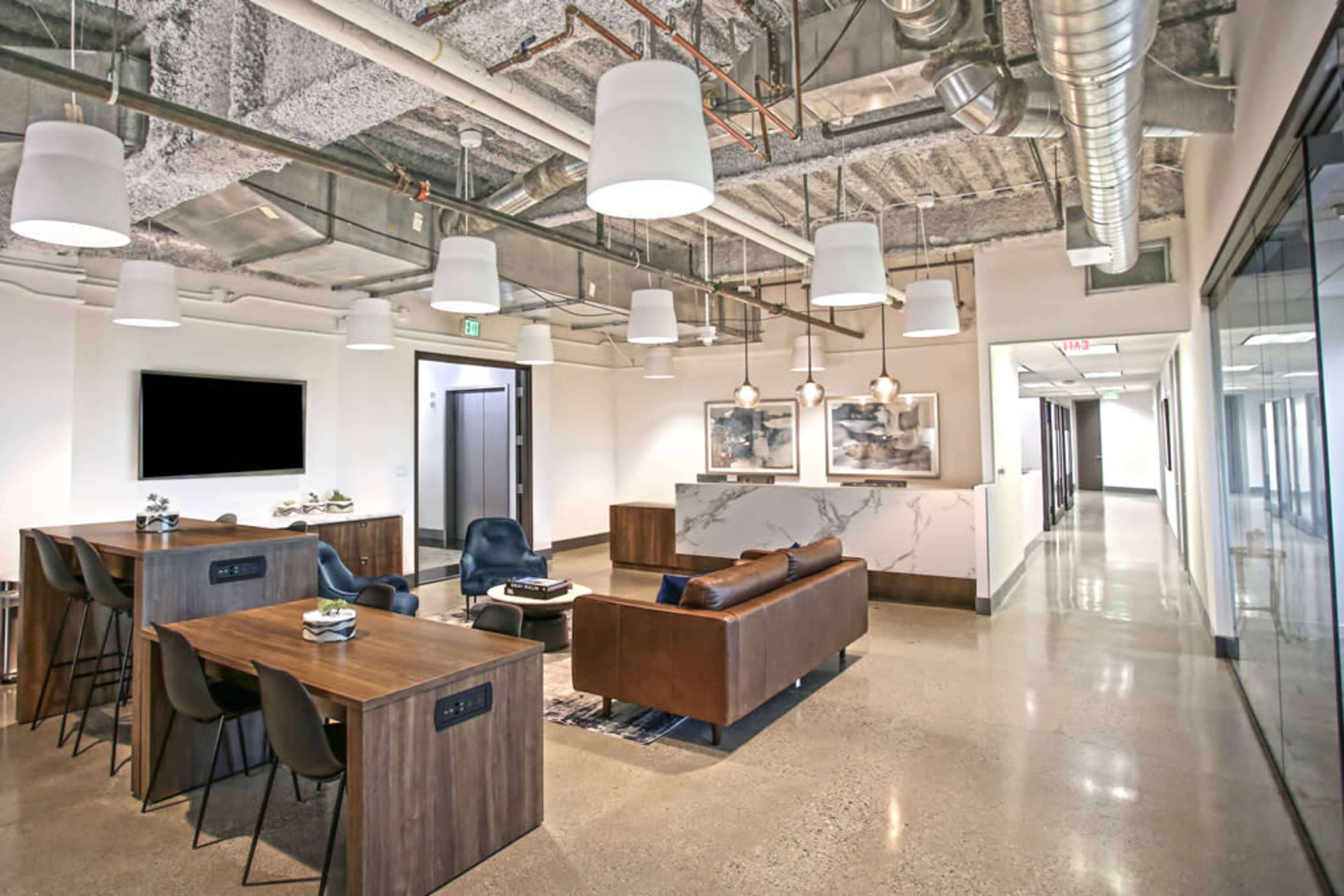 The image shows a modern office lounge area featuring a seating arrangement with a sofa and chairs, several tables, and exposed ceiling ducts.