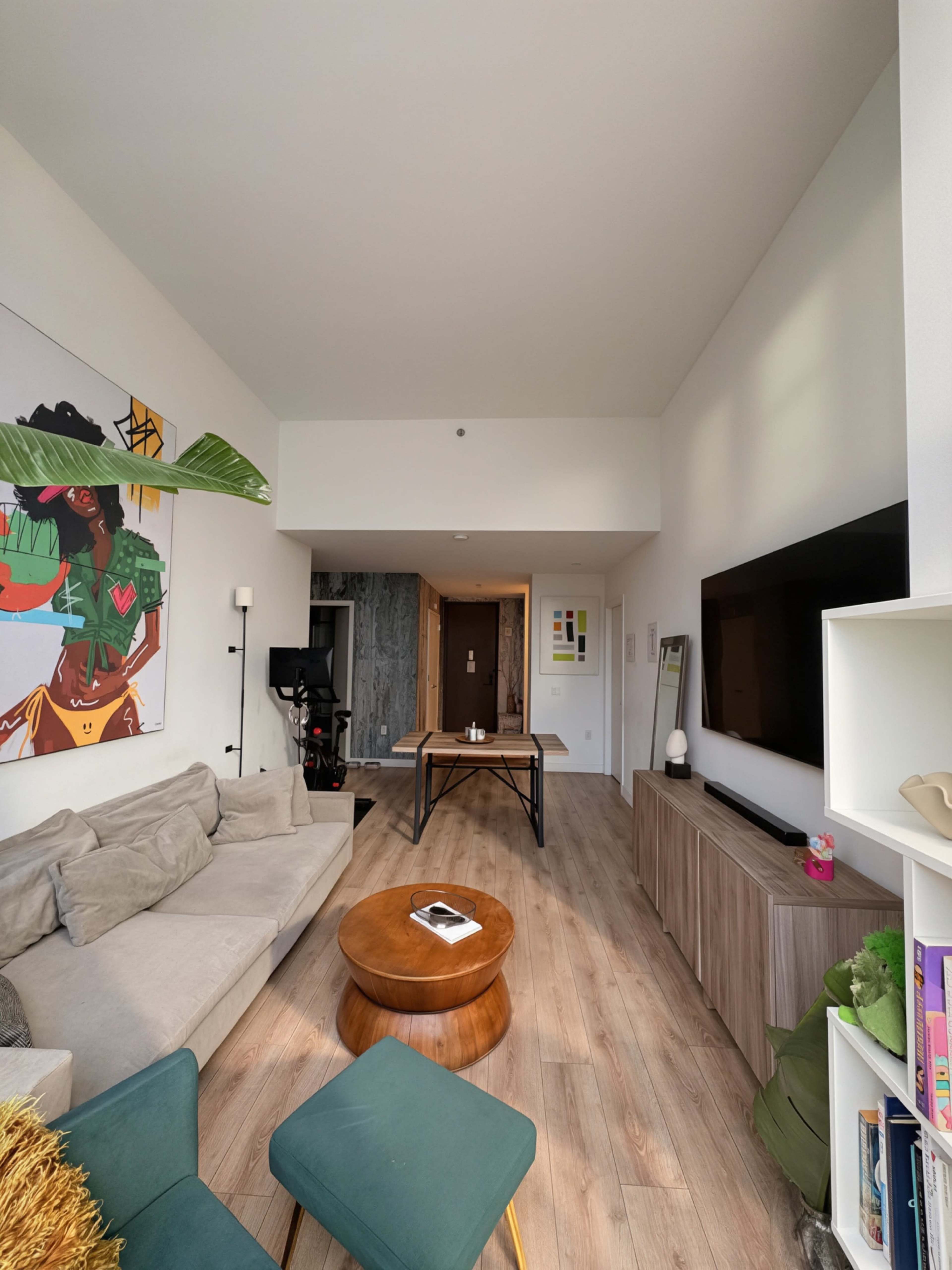 The image shows a modern living room with a beige sofa, a round wooden coffee table, and a wall-mounted television, featuring colorful artwork and a doorway leading to another room.
