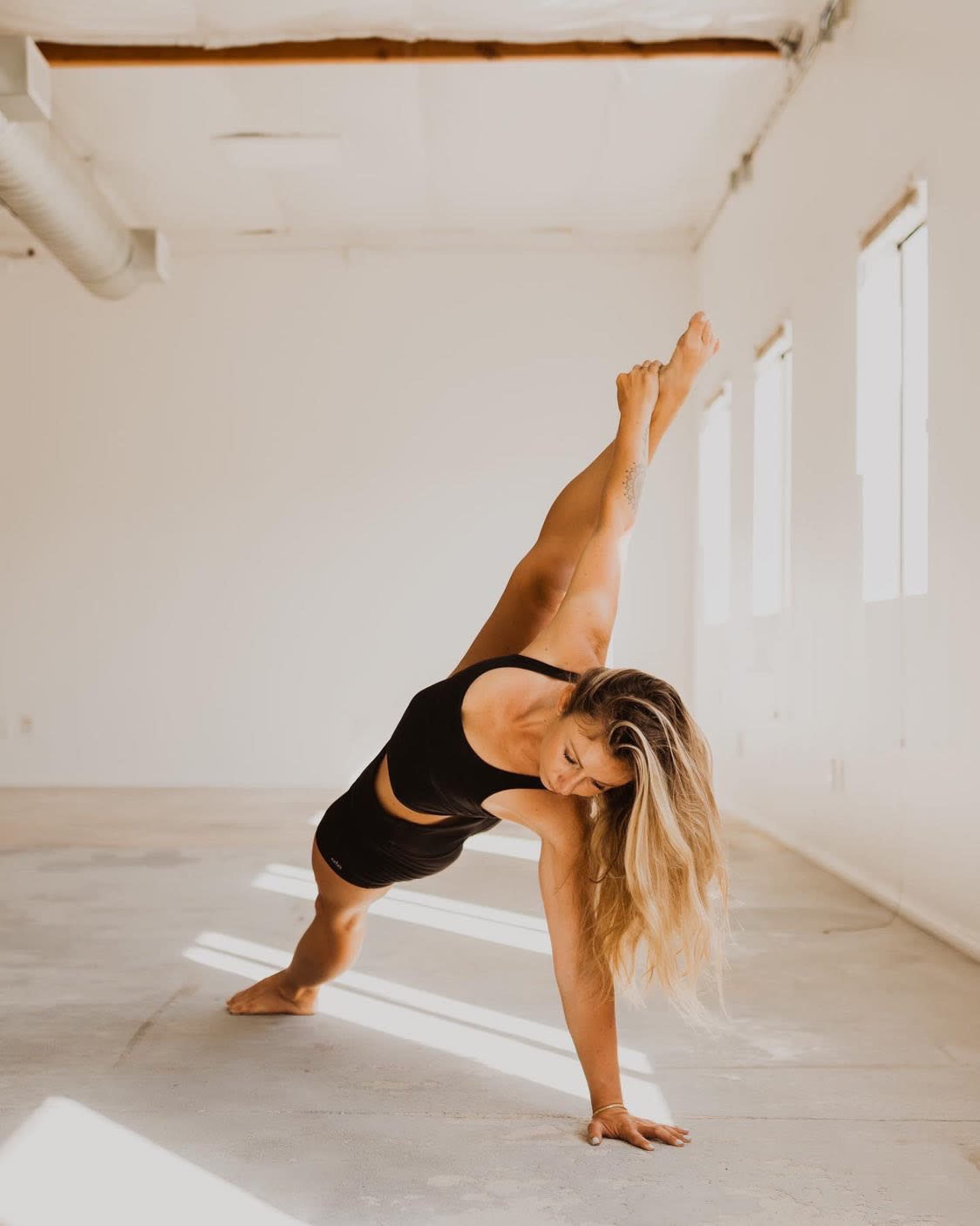 A woman performs a side plank yoga pose on a light-colored floor in a minimalist room with natural light.