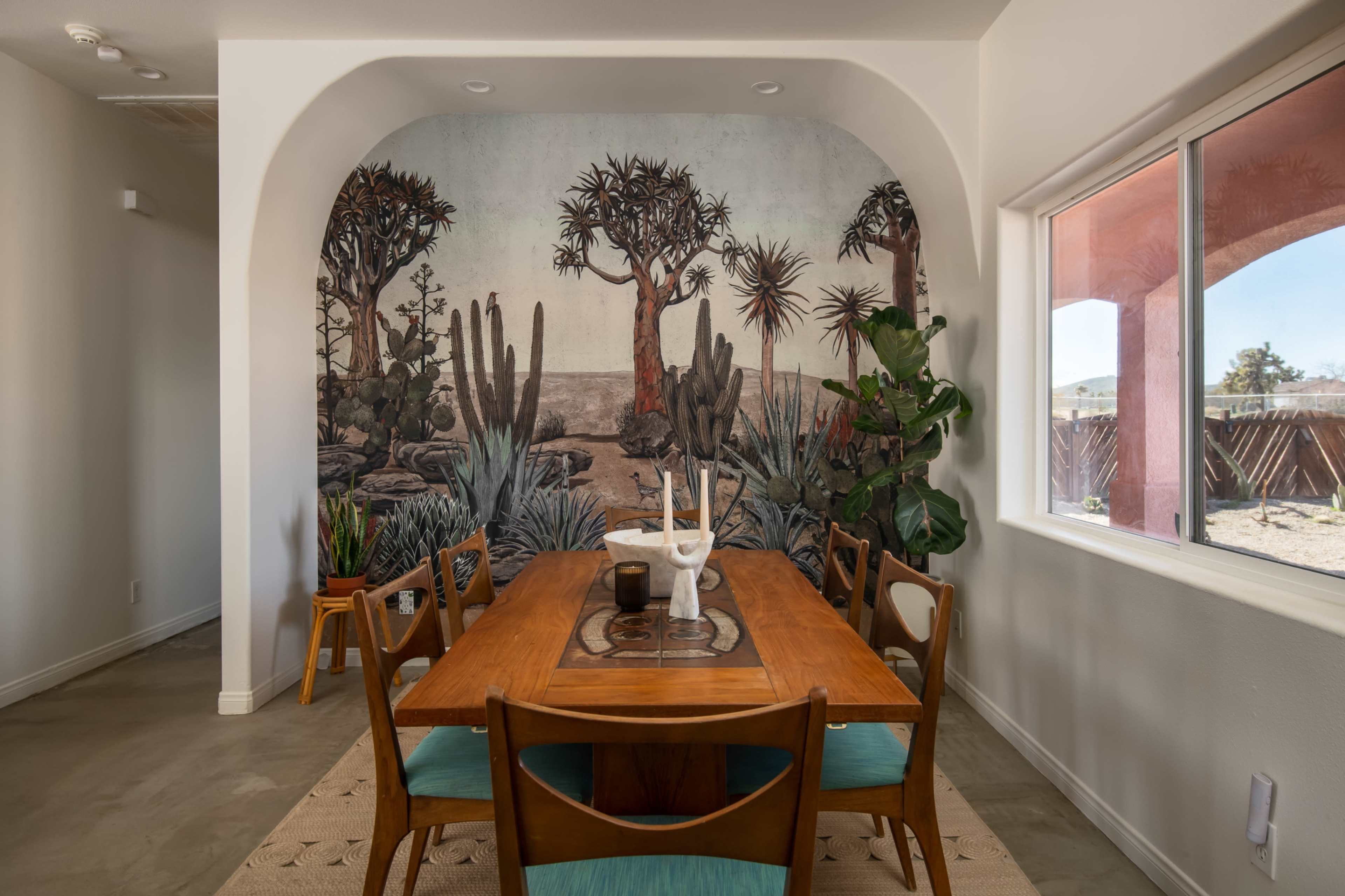 A dining room features a wooden table surrounded by chairs, with a mural of desert plants and trees on the wall.