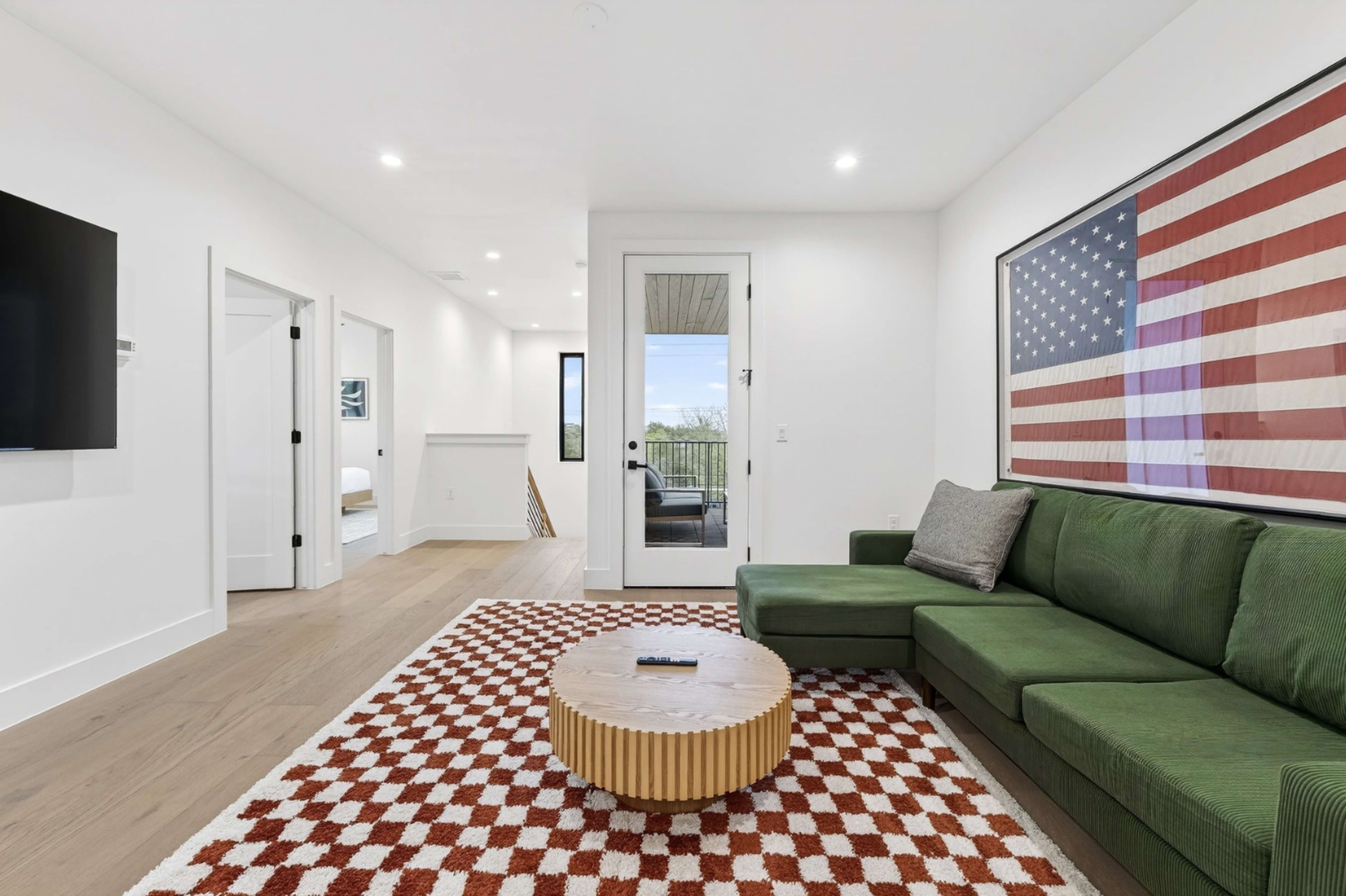 The image shows a modern living room featuring a green sectional sofa, a round wooden coffee table, a red and white patterned rug, and large windows with a view.