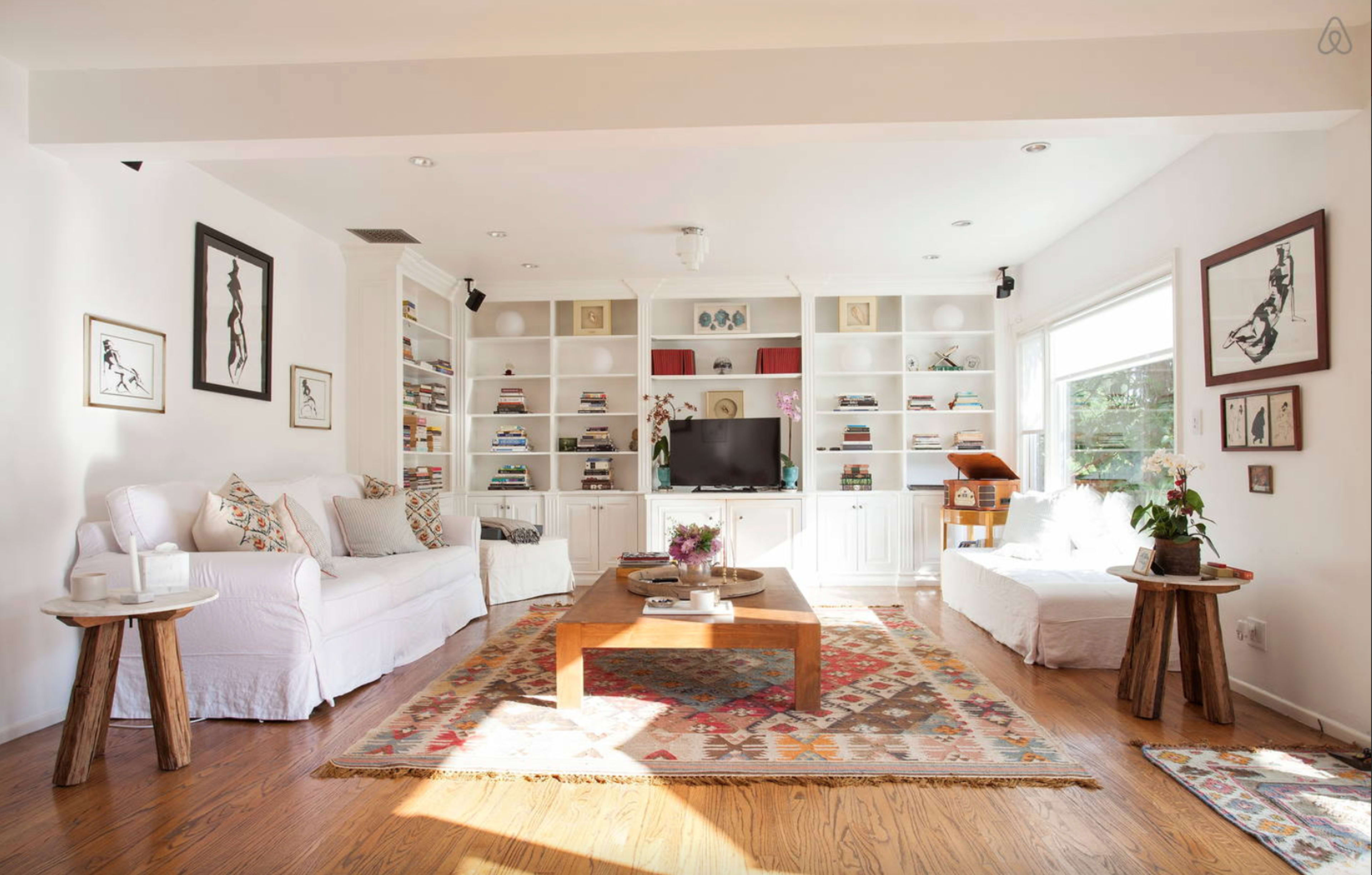 The image shows a bright living room with two white sofas, a wooden coffee table, and built-in shelves filled with books and decor.