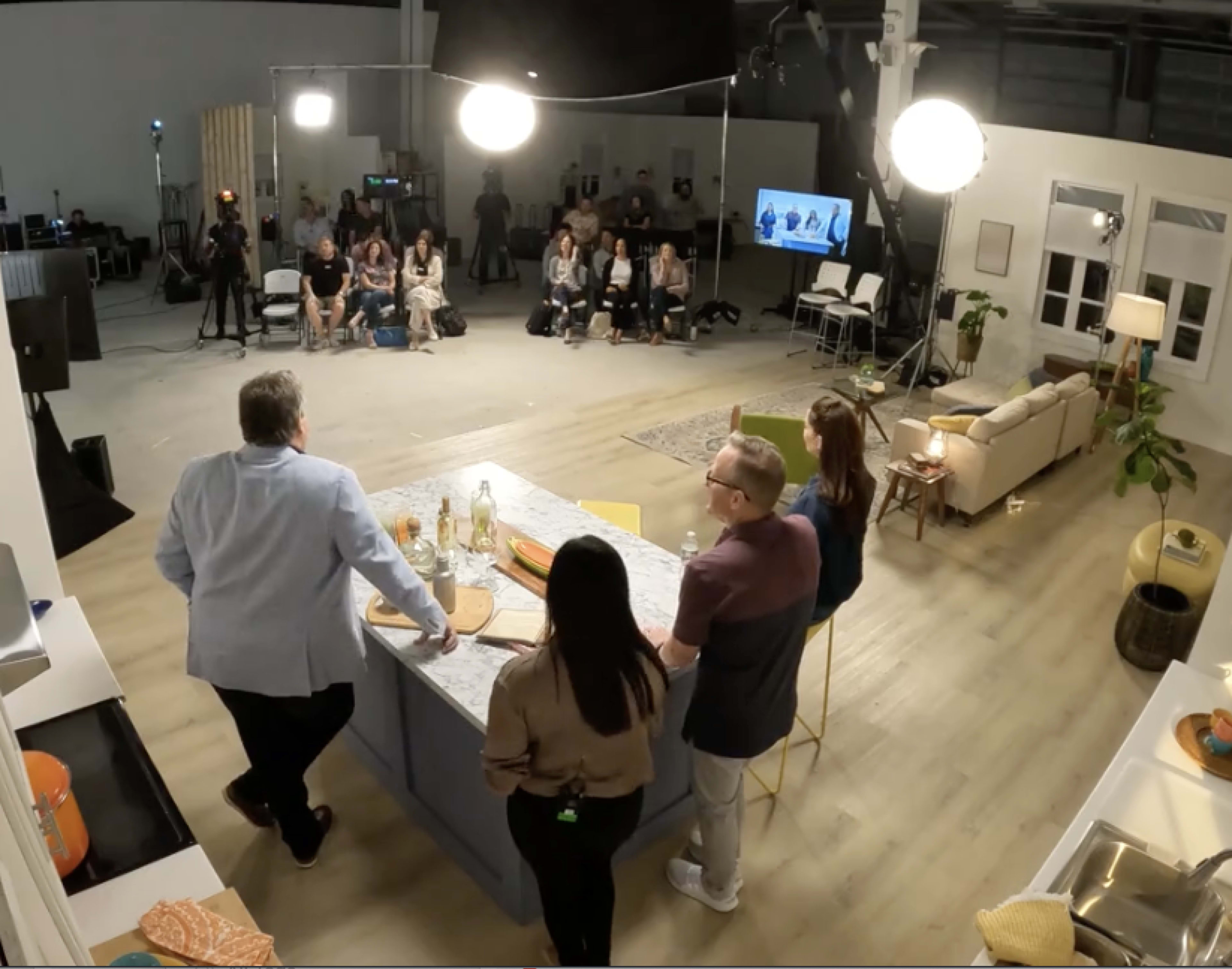 A group of people is gathered in a studio setting, with a kitchen island in the foreground and an audience seated in the background.