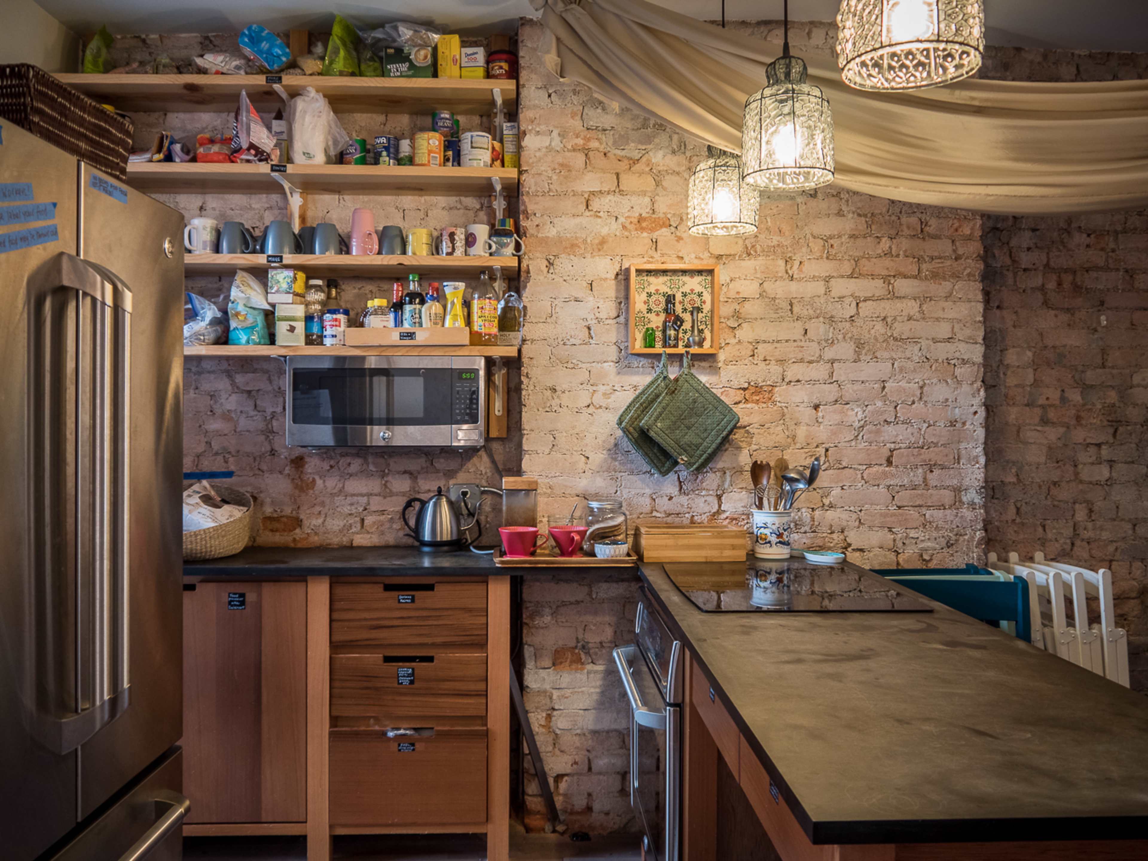 The kitchen features exposed brick walls, open shelving filled with various kitchen items, a stainless steel refrigerator, and a countertop with utensils and appliances.