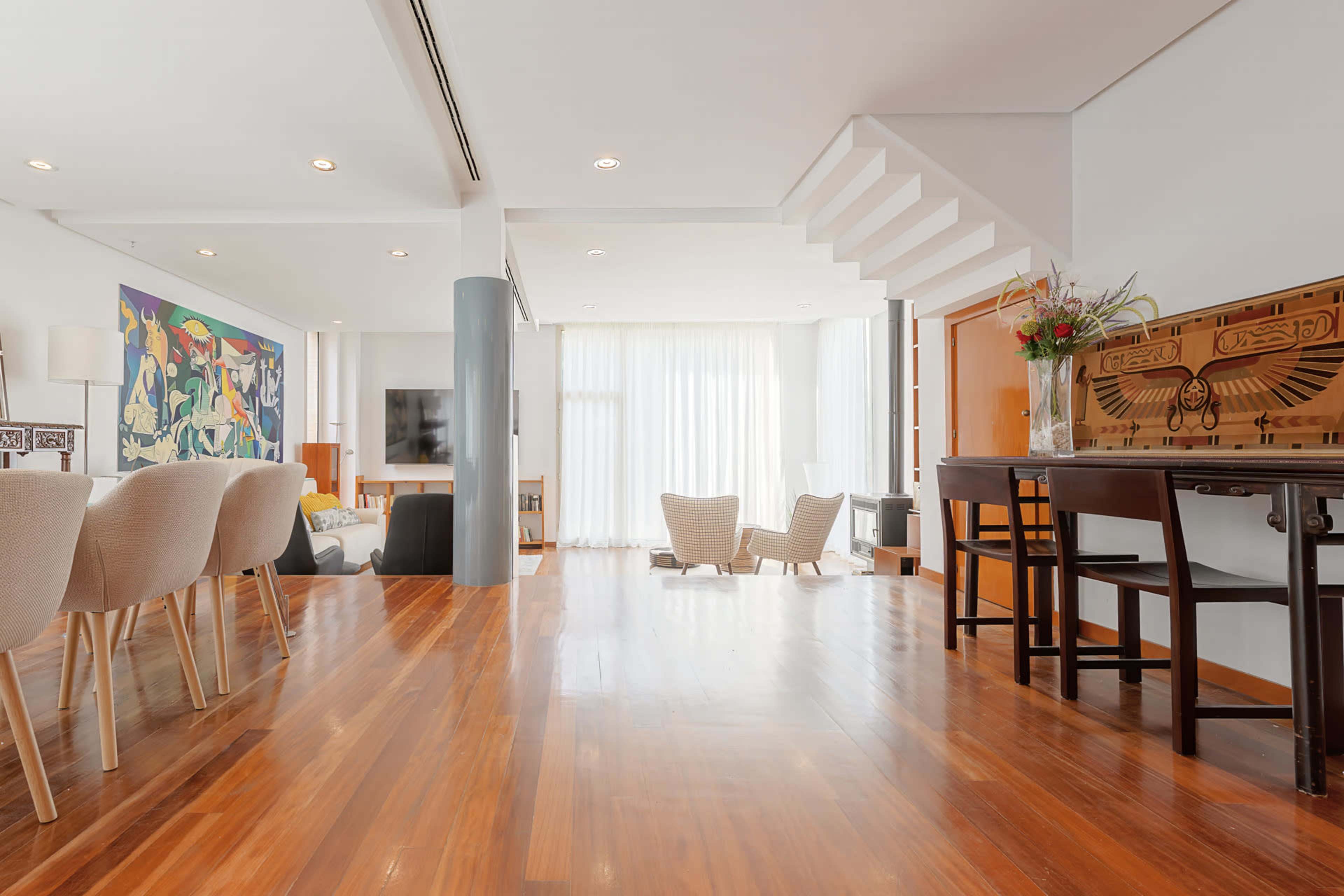 The image shows a spacious living area with wooden floors, modern furniture, a dining table, and large windows allowing natural light to enter.