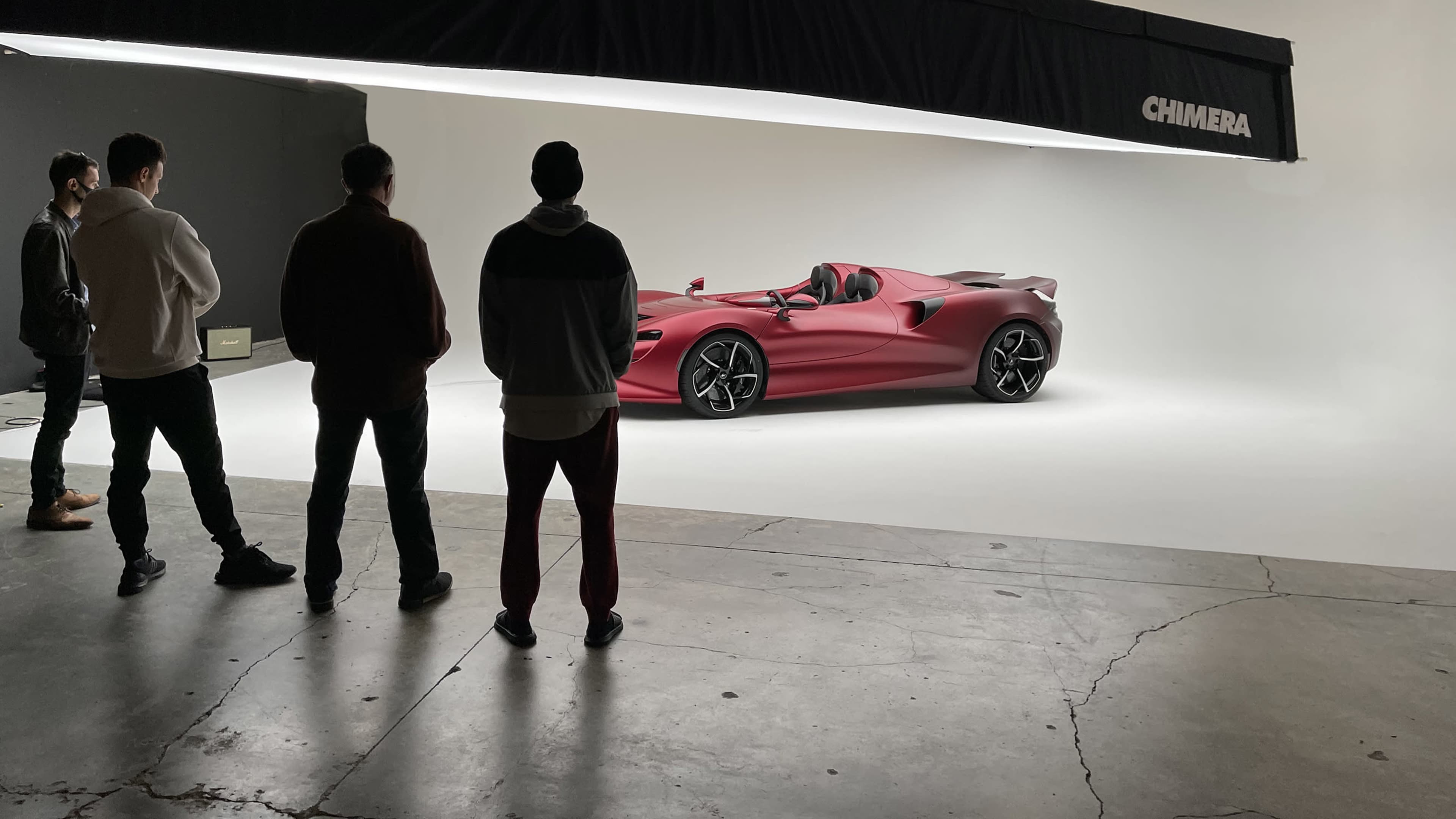 A group of five people stands in a studio, observing a sleek red sports car positioned on a white backdrop.