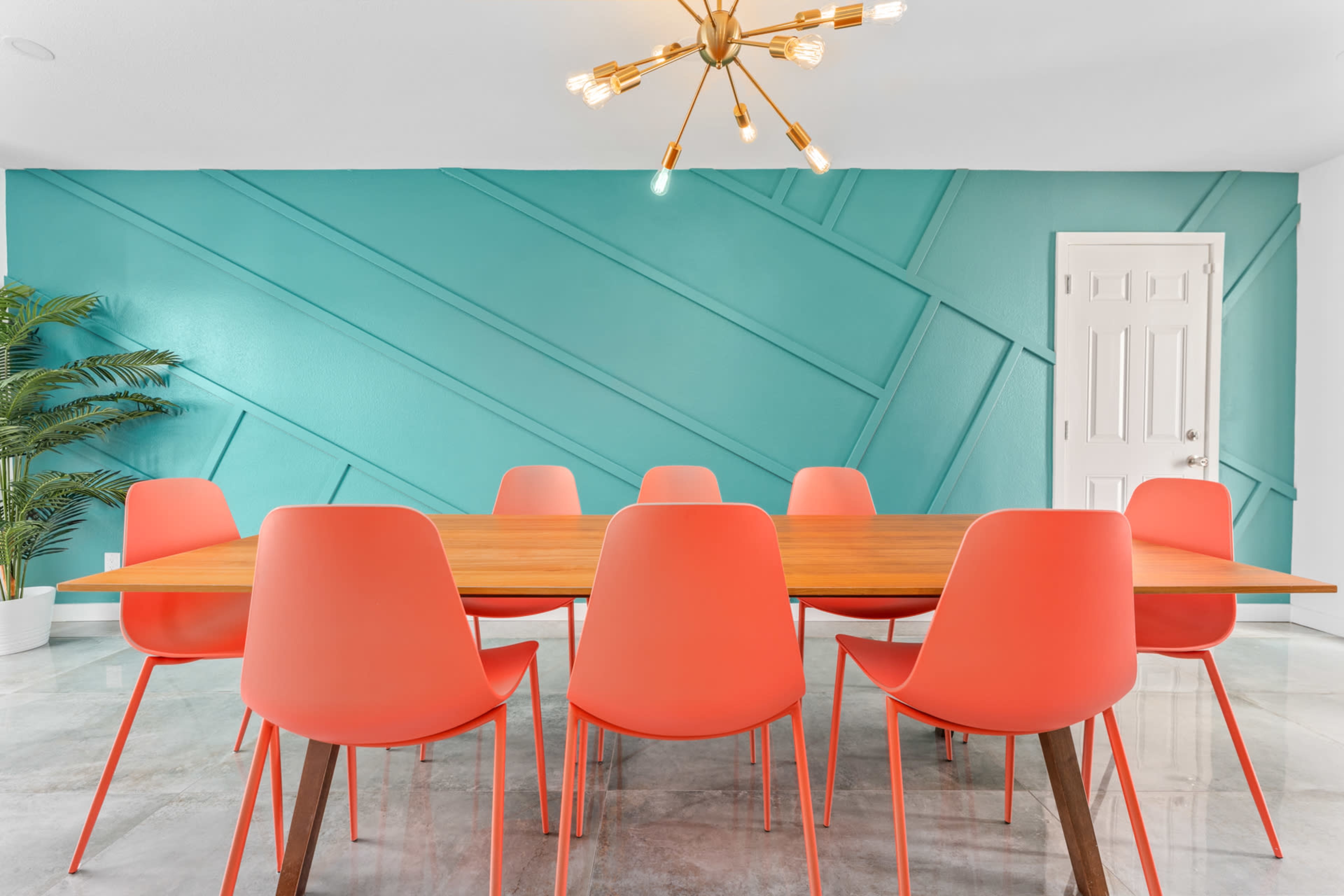 A modern dining room features a large wooden table surrounded by eight coral-colored chairs against a turquoise accent wall.