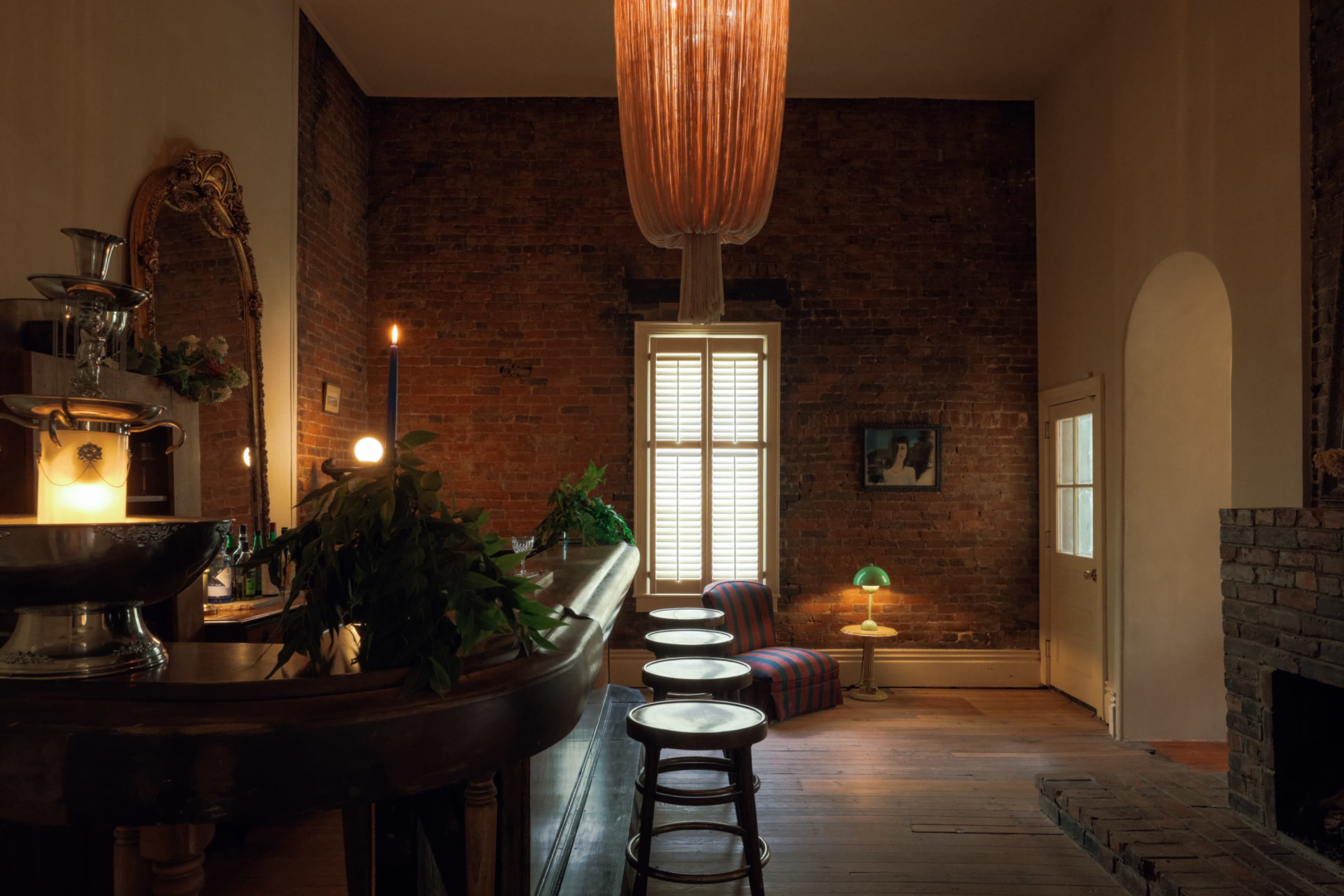 A bar area with wooden stools and a vibrant hanging light fixture is set against exposed brick walls, next to a window with shutters and a cozy seating arrangement.