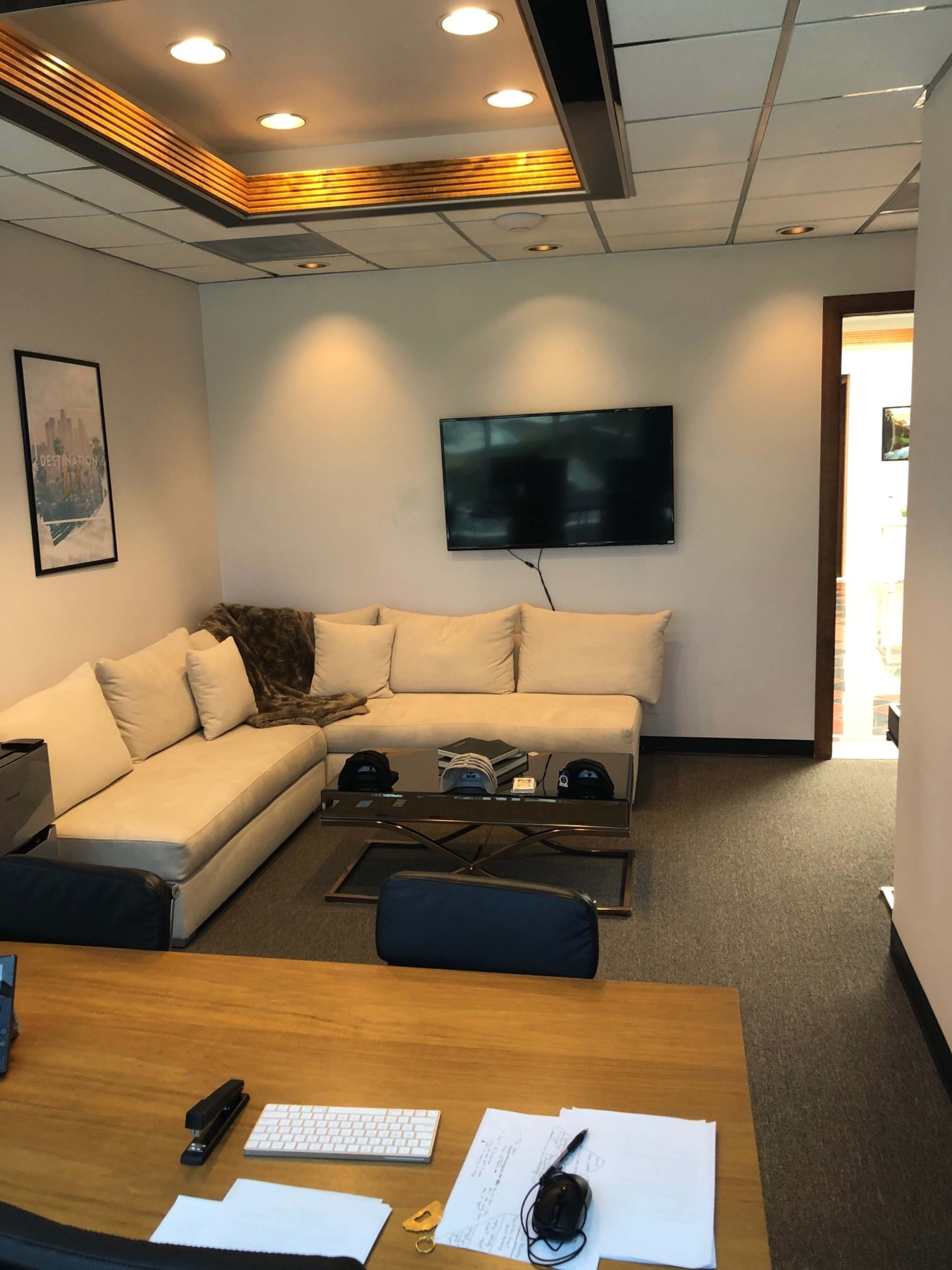 A modern office space featuring a light-colored sectional sofa, a wall-mounted television, and a wooden desk with documents and a telephone on it.
