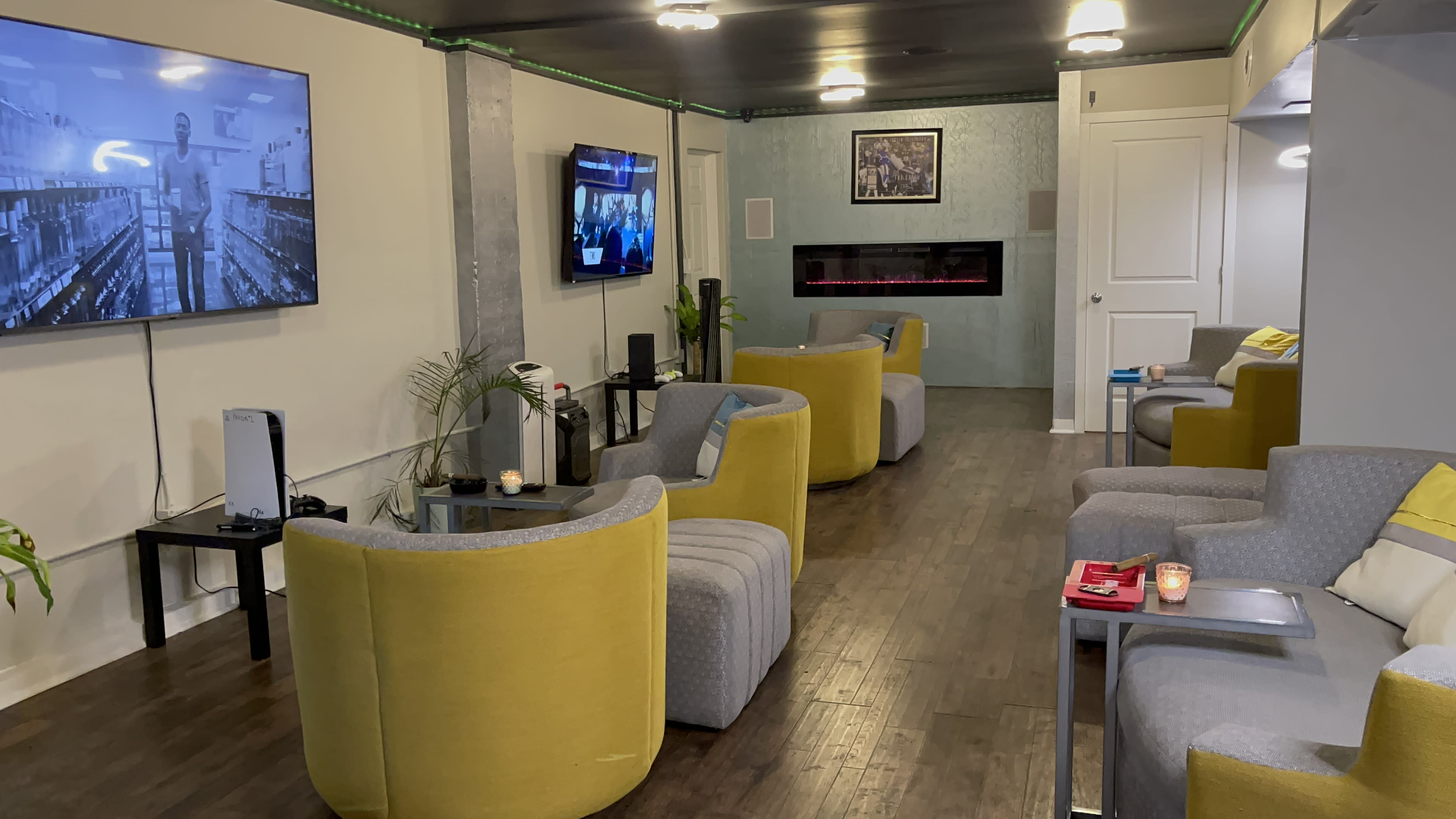 A modern waiting area with yellow and gray armchairs, two televisions, a faux fireplace, and a plant.