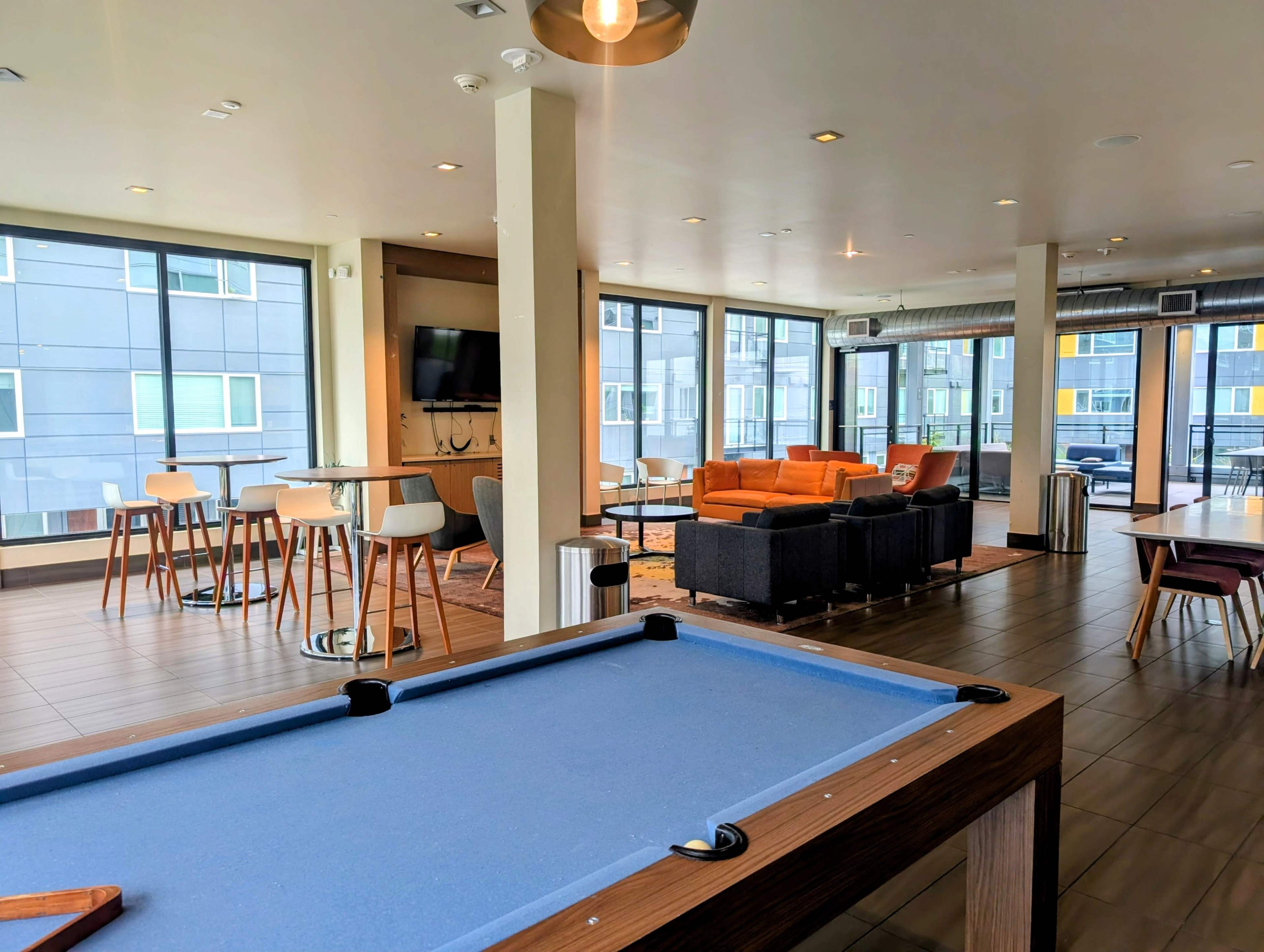 The image shows a common area with a billiard table in the foreground and seating arrangements in a well-lit space featuring large windows.