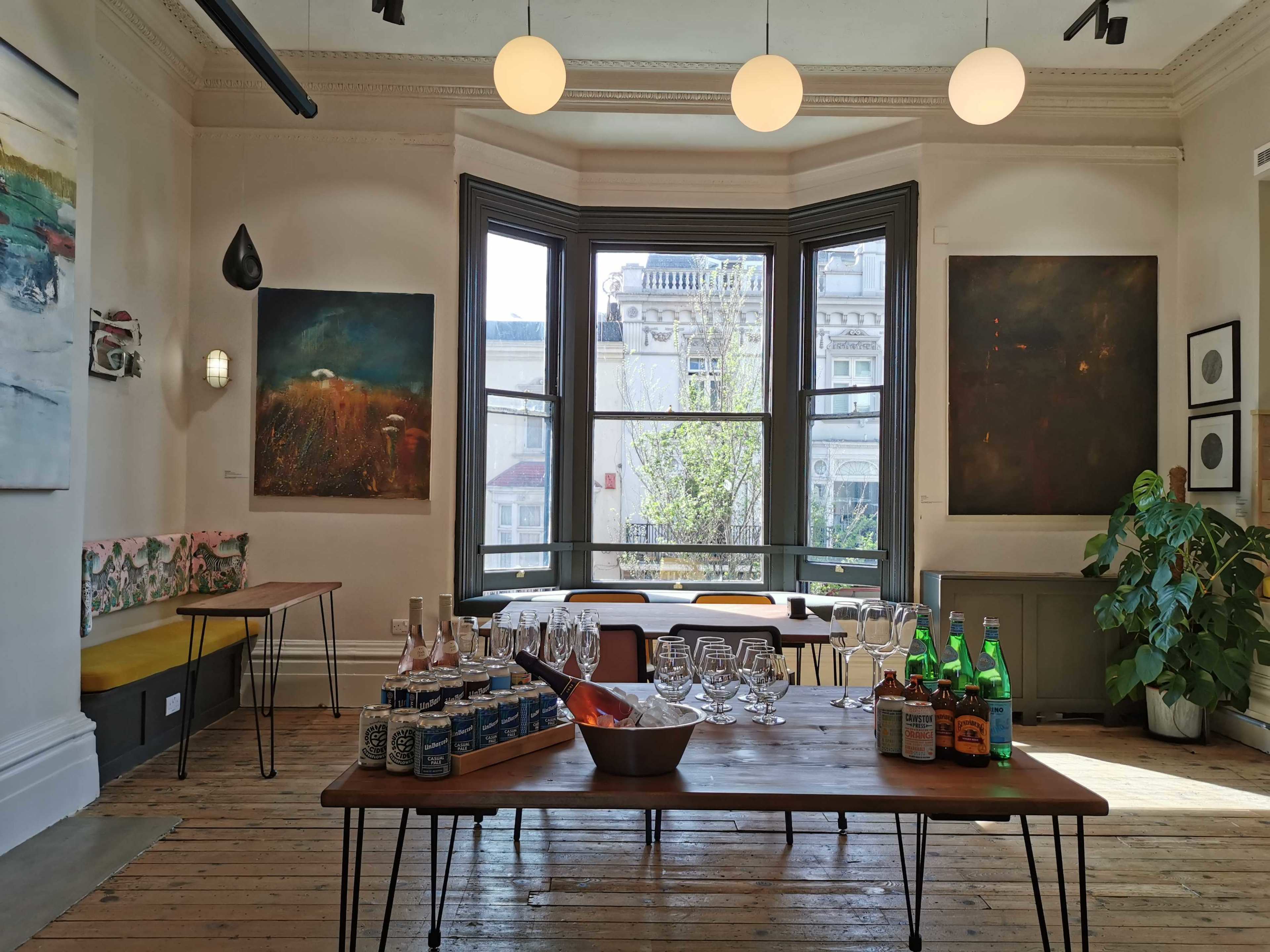 An art gallery with large windows, a table set with bottles and glasses, and artwork displayed on the walls.