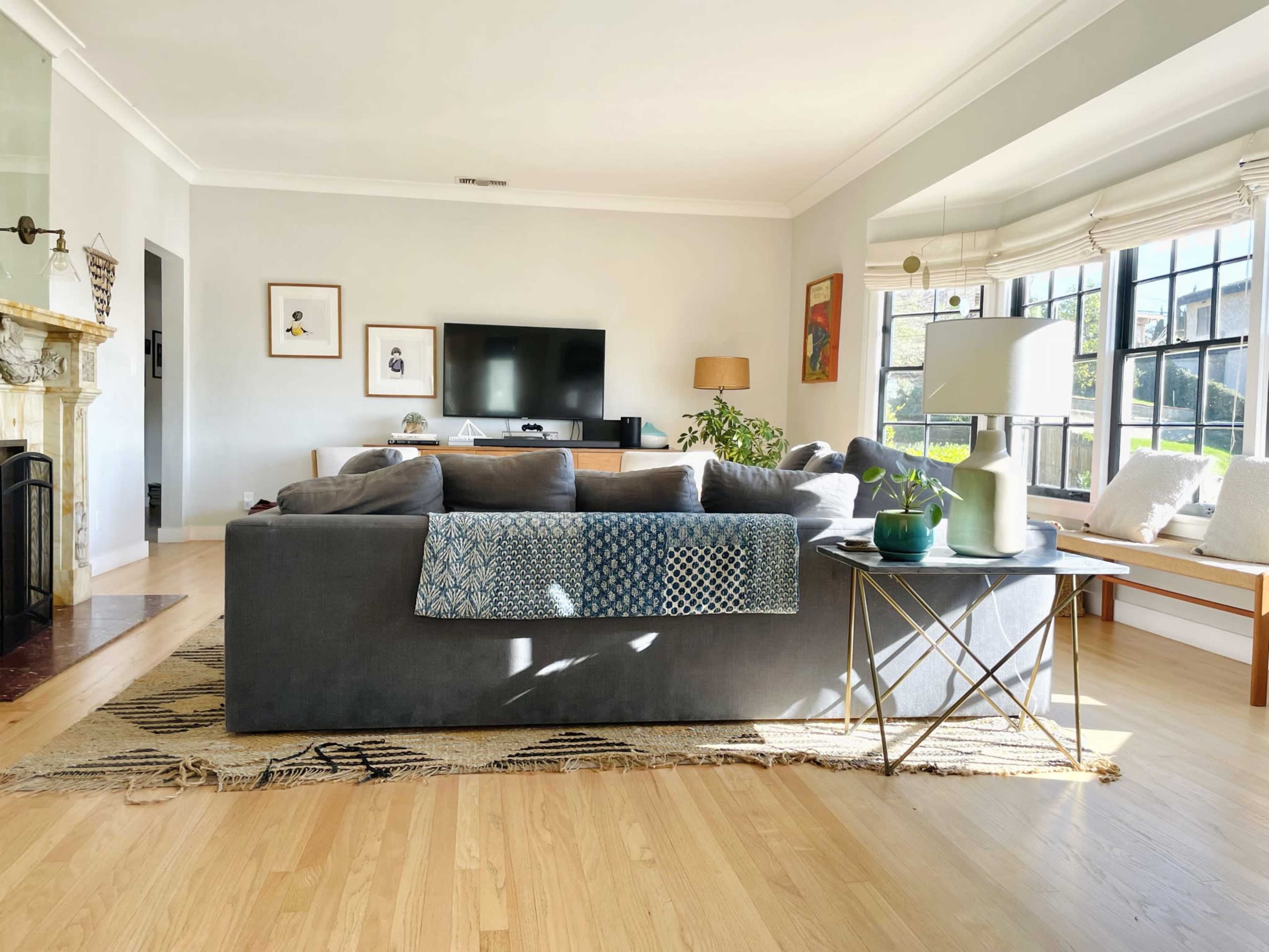 The living room features a gray sectional sofa with a throw blanket, a television stand, and large windows allowing natural light to enter.