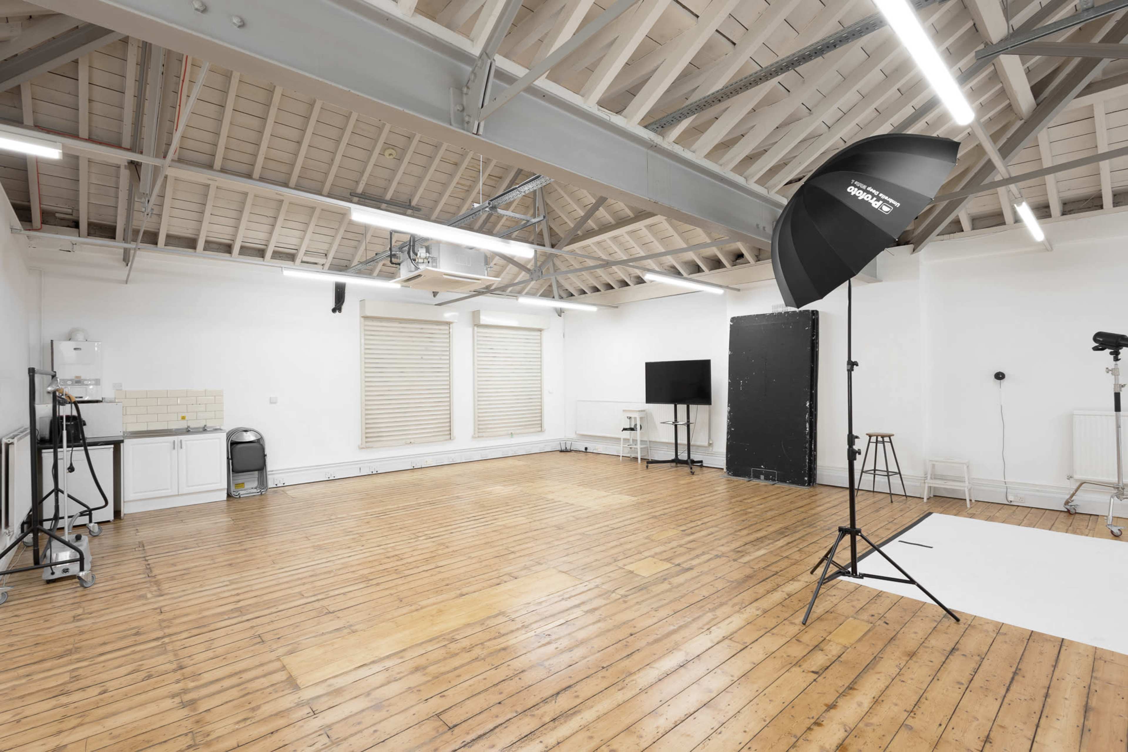 The image shows a spacious, well-lit studio with wooden flooring, a backdrop stand, a light stand with a softbox, and a small kitchenette area.