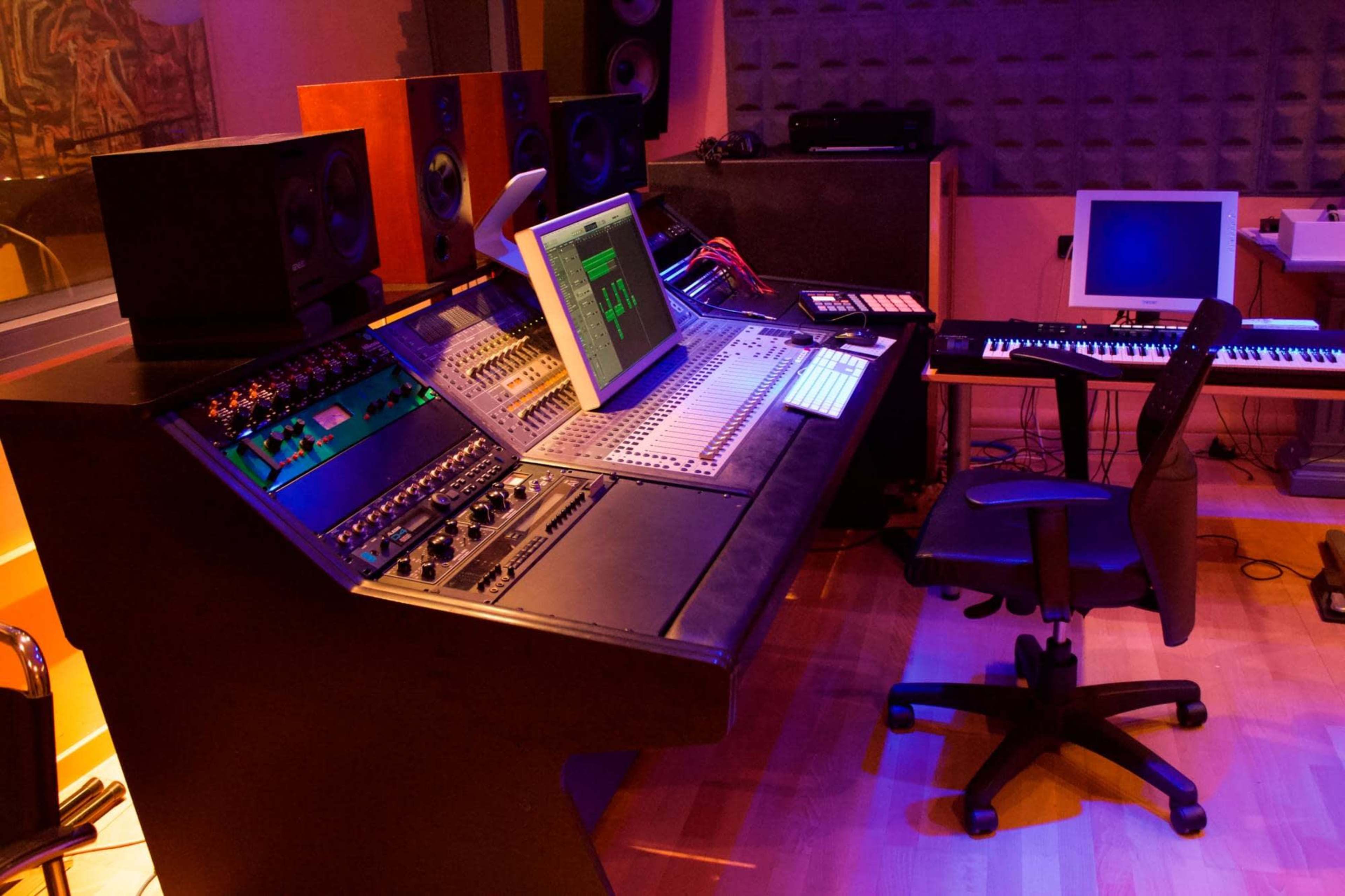 A music studio featuring a large mixing console, computer monitors, speakers, and a keyboard arranged around a workspace.