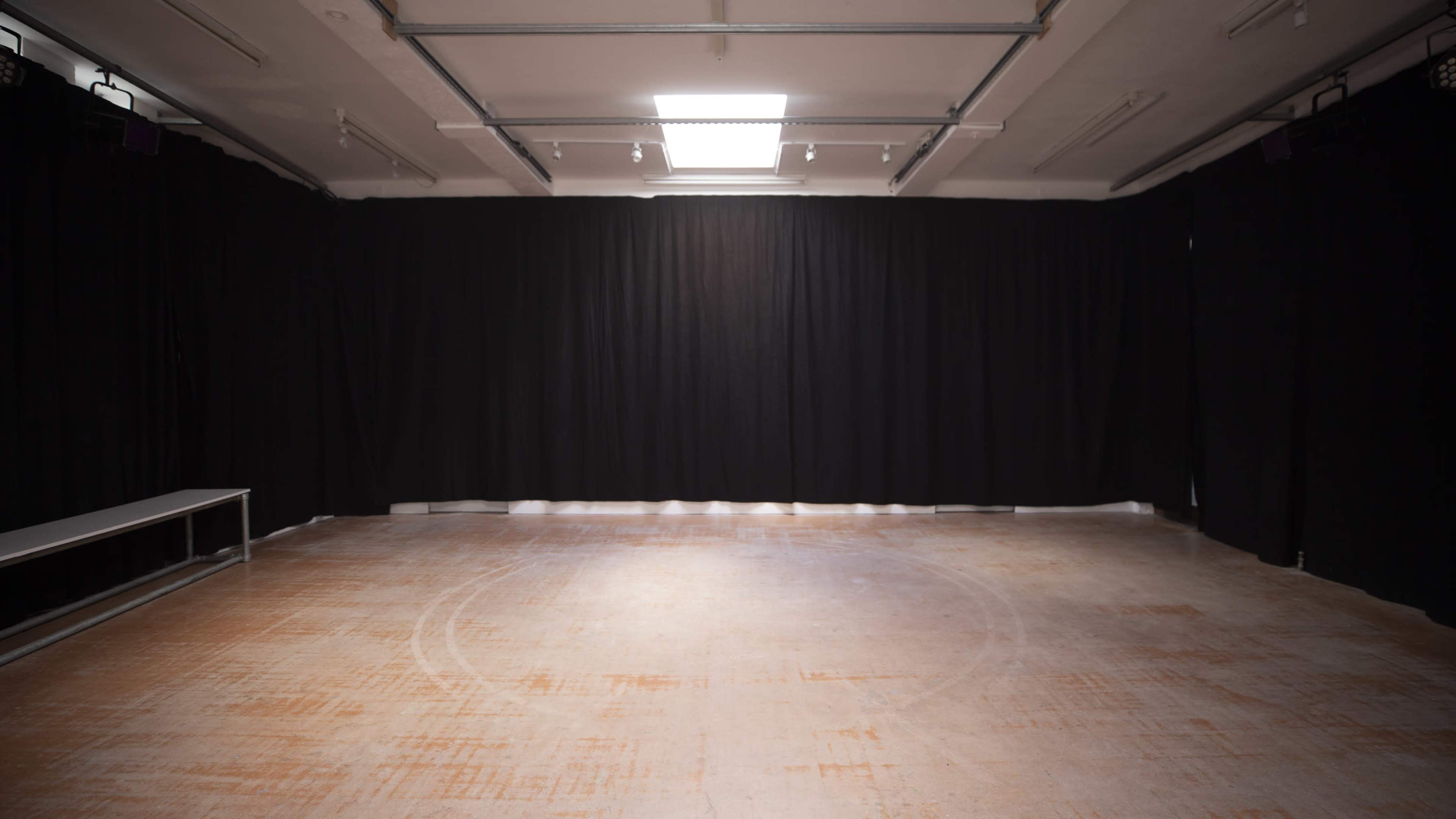 An empty room with a wooden floor and black curtains lining the walls, illuminated by natural light from a ceiling skylight.