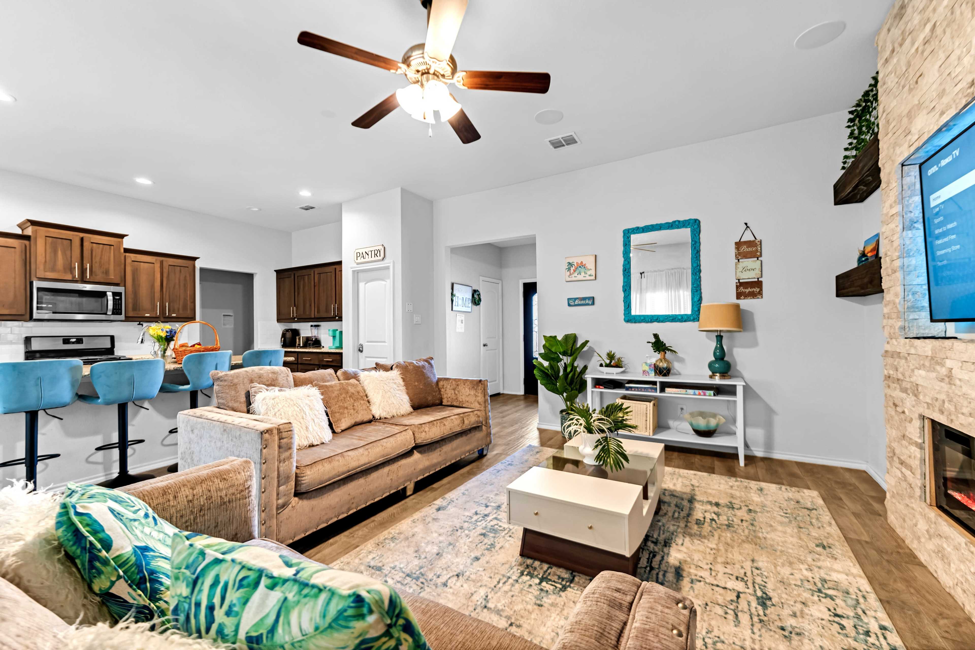 The living room features a sofa and armchair with a coffee table, a modern kitchen area in the background, and a stone fireplace beneath a mounted television.