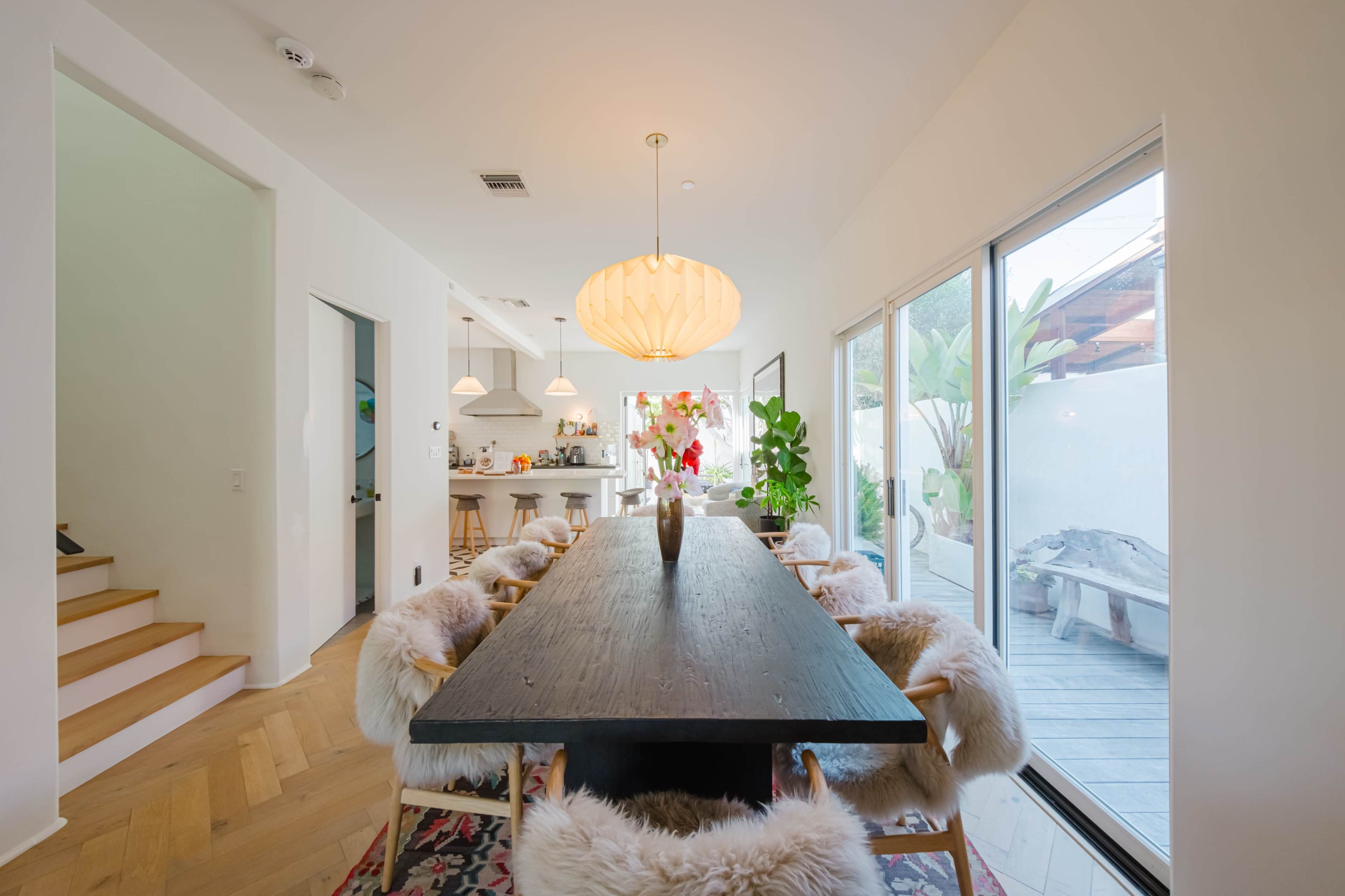 A long wooden dining table surrounded by chairs with fur cushions is positioned in a bright room that leads to an outdoor space.