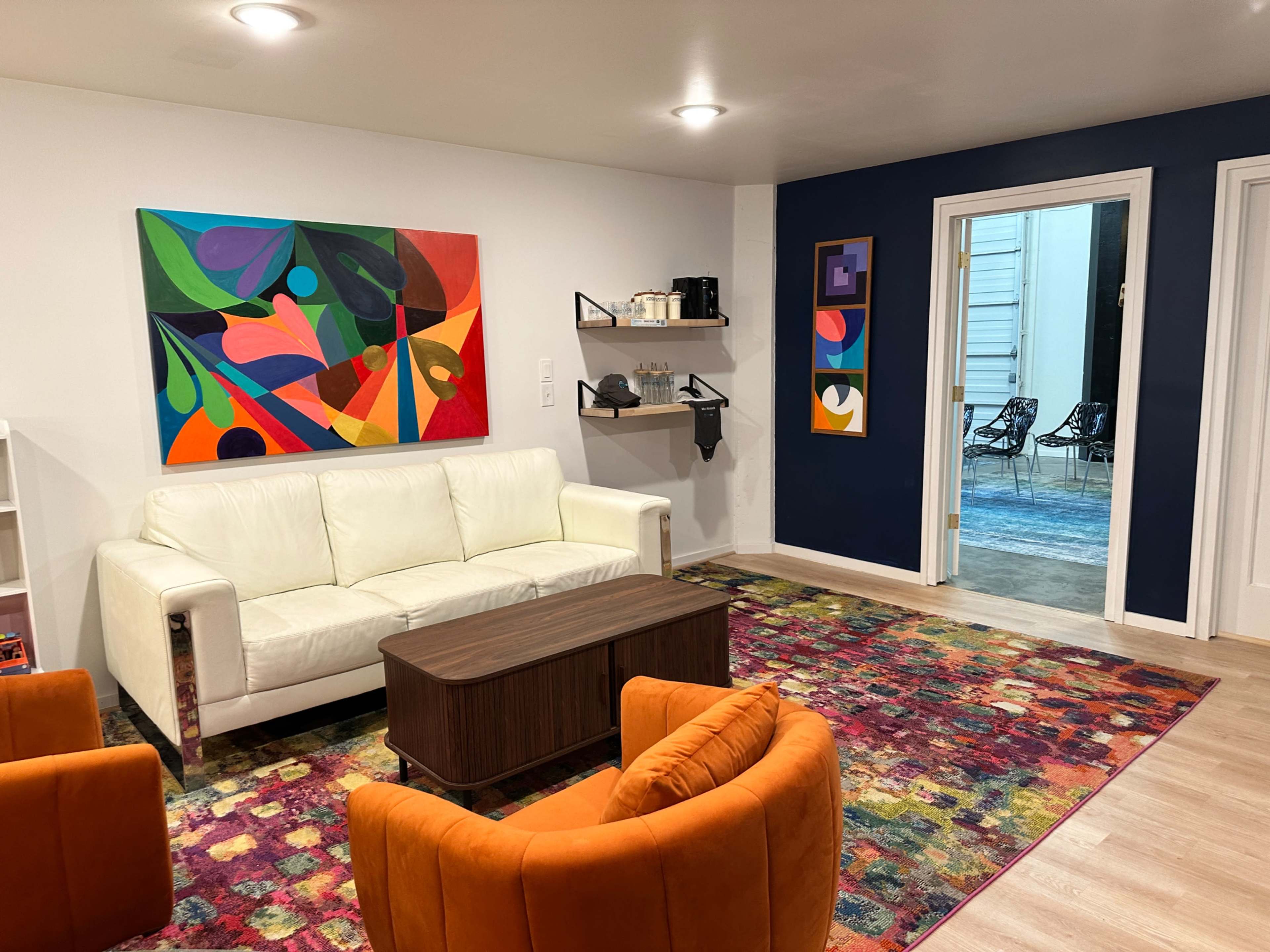 The image shows a modern living room with a white sofa, a colorful rug, and artwork on the walls, featuring a coffee table and shelves with decorative items.