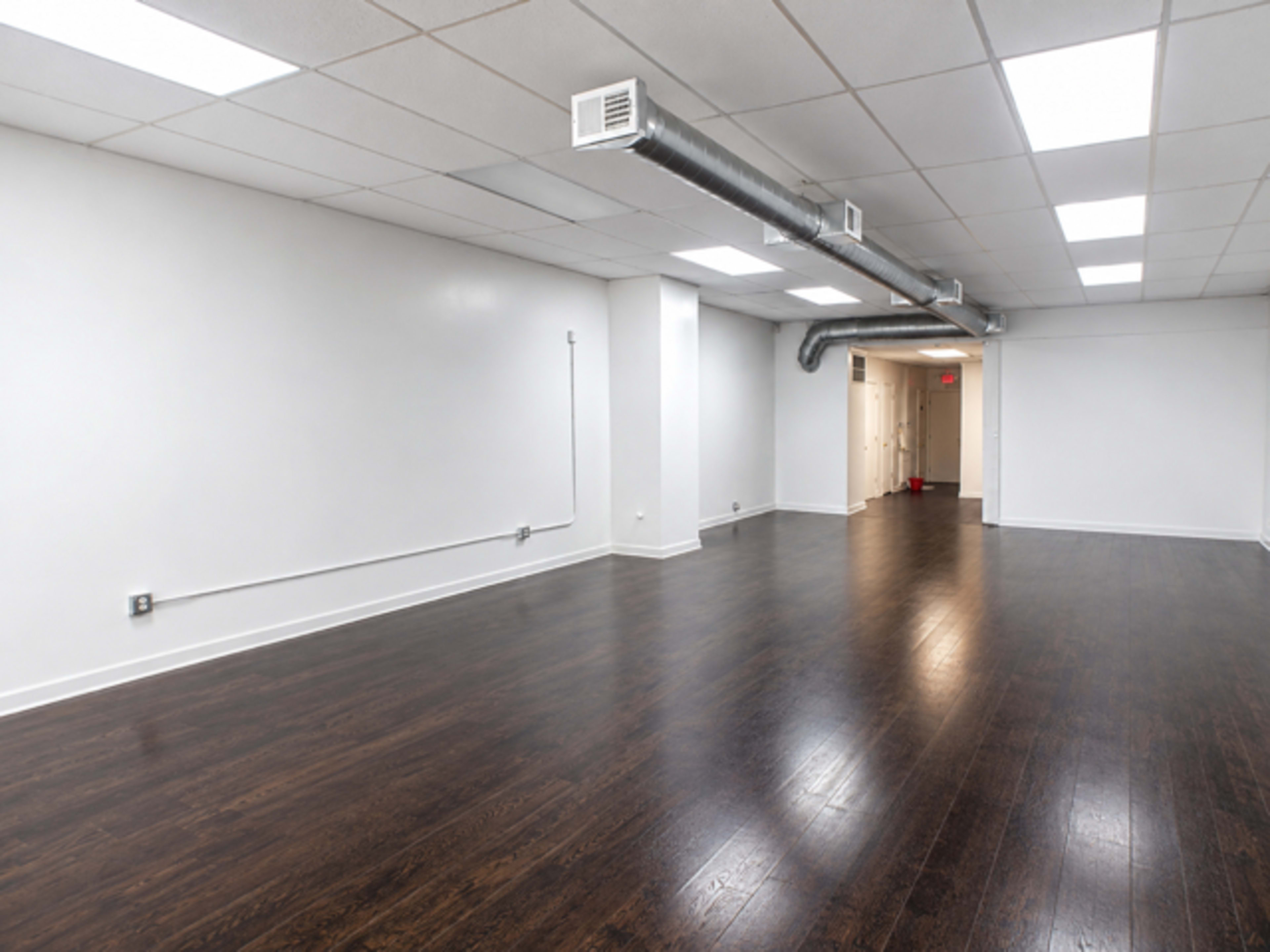 The image shows a spacious, empty room with dark wood flooring and white walls, featuring ductwork on the ceiling and a doorway at the back.
