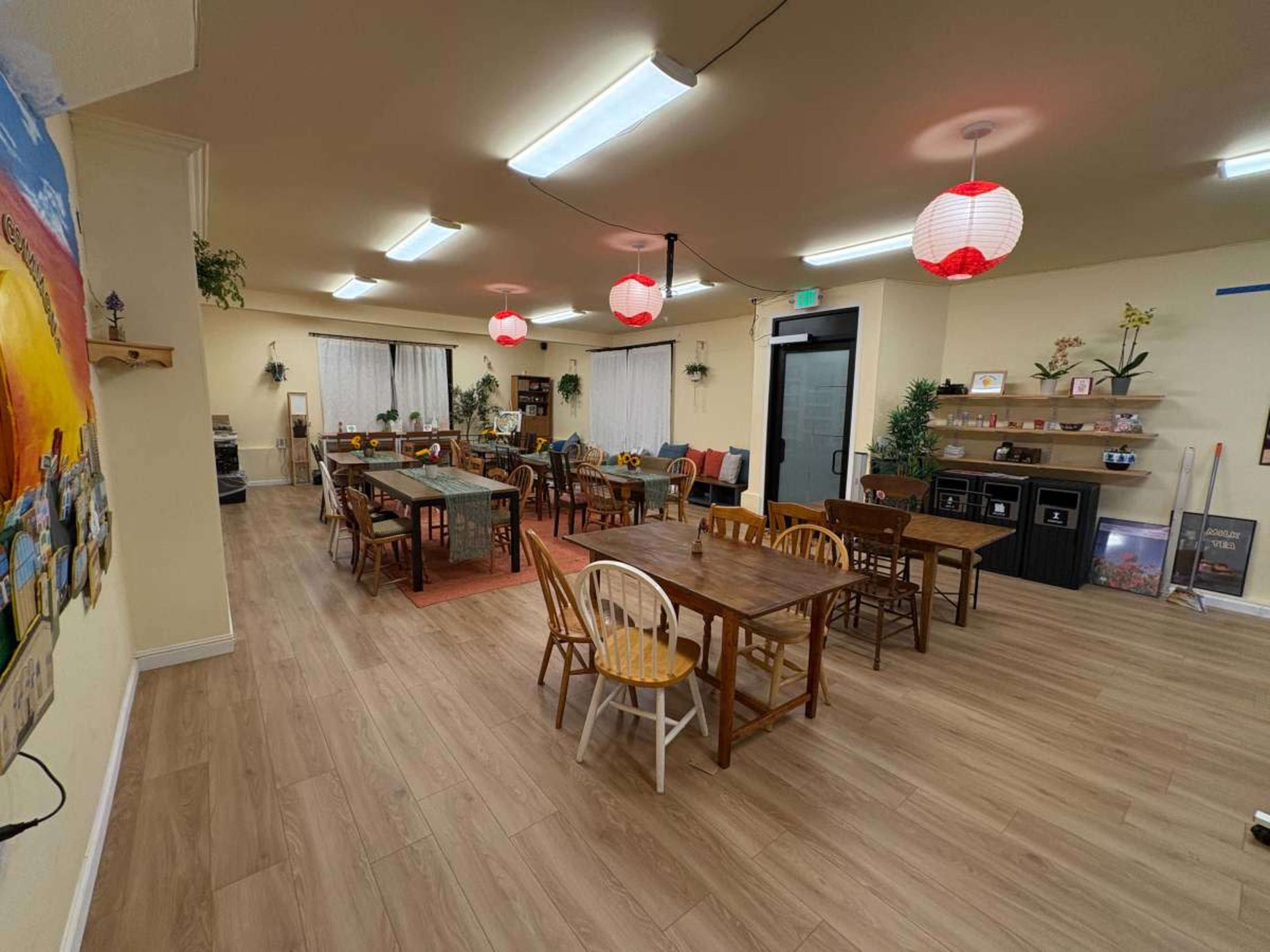 The image depicts a spacious room with multiple wooden tables and chairs arranged neatly, along with decorative elements and soft lighting.