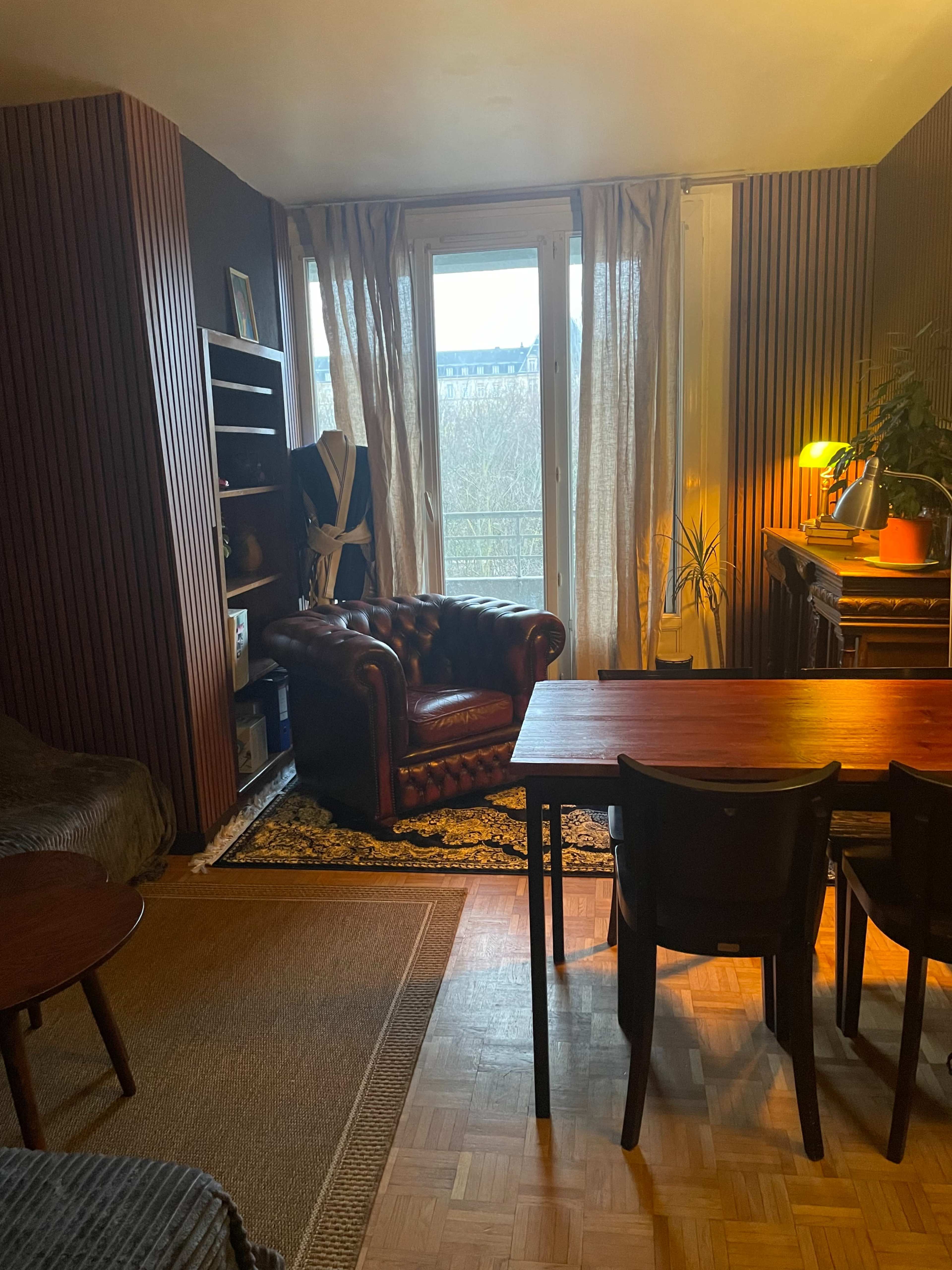 The image shows a cozy living space with a leather armchair, a wooden dining table with chairs, and shelves against a wall, illuminated by natural light from large windows.
