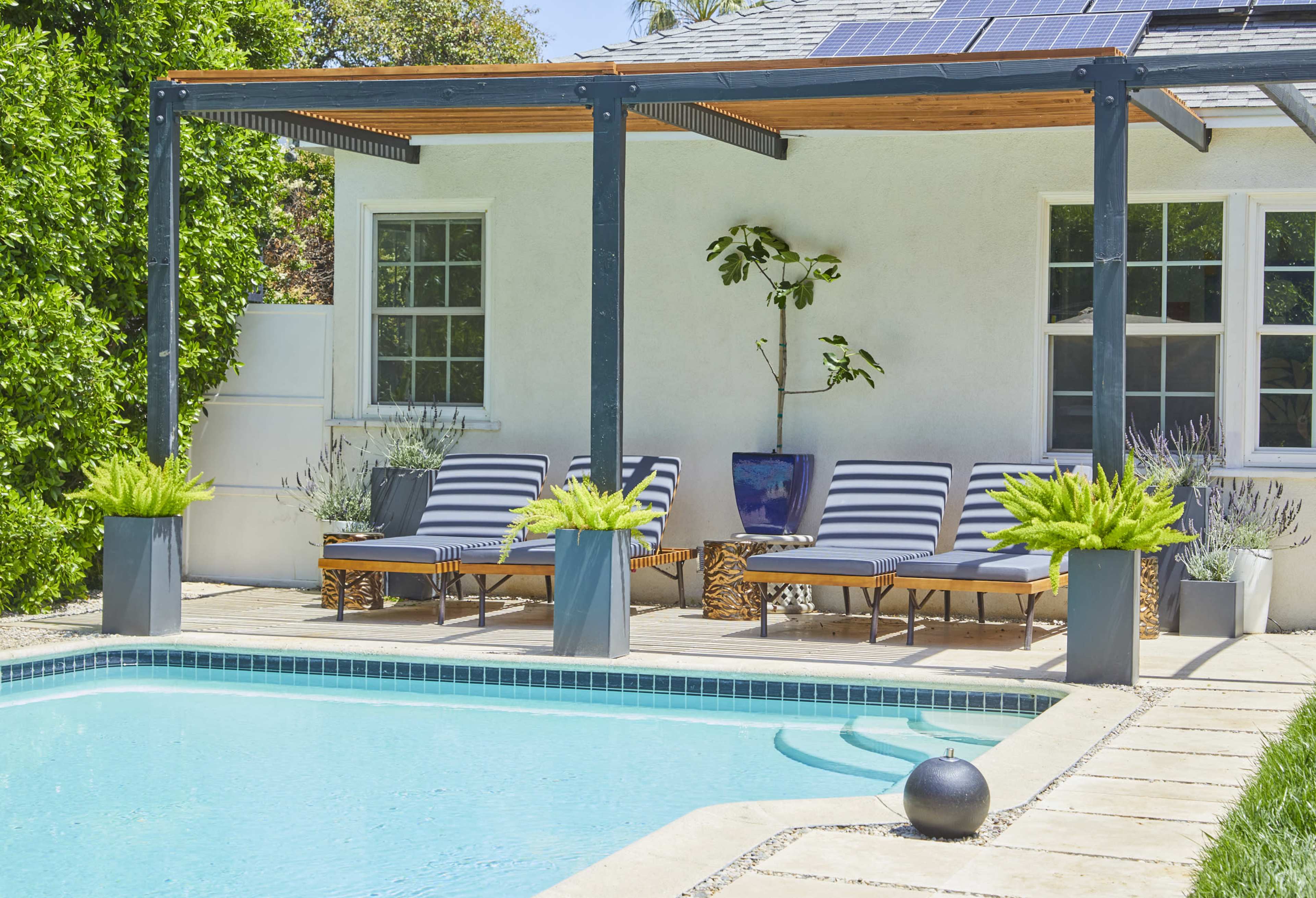 The image shows a serene patio area with two striped lounge chairs under a pergola near a swimming pool, surrounded by greenery and decorative planters.