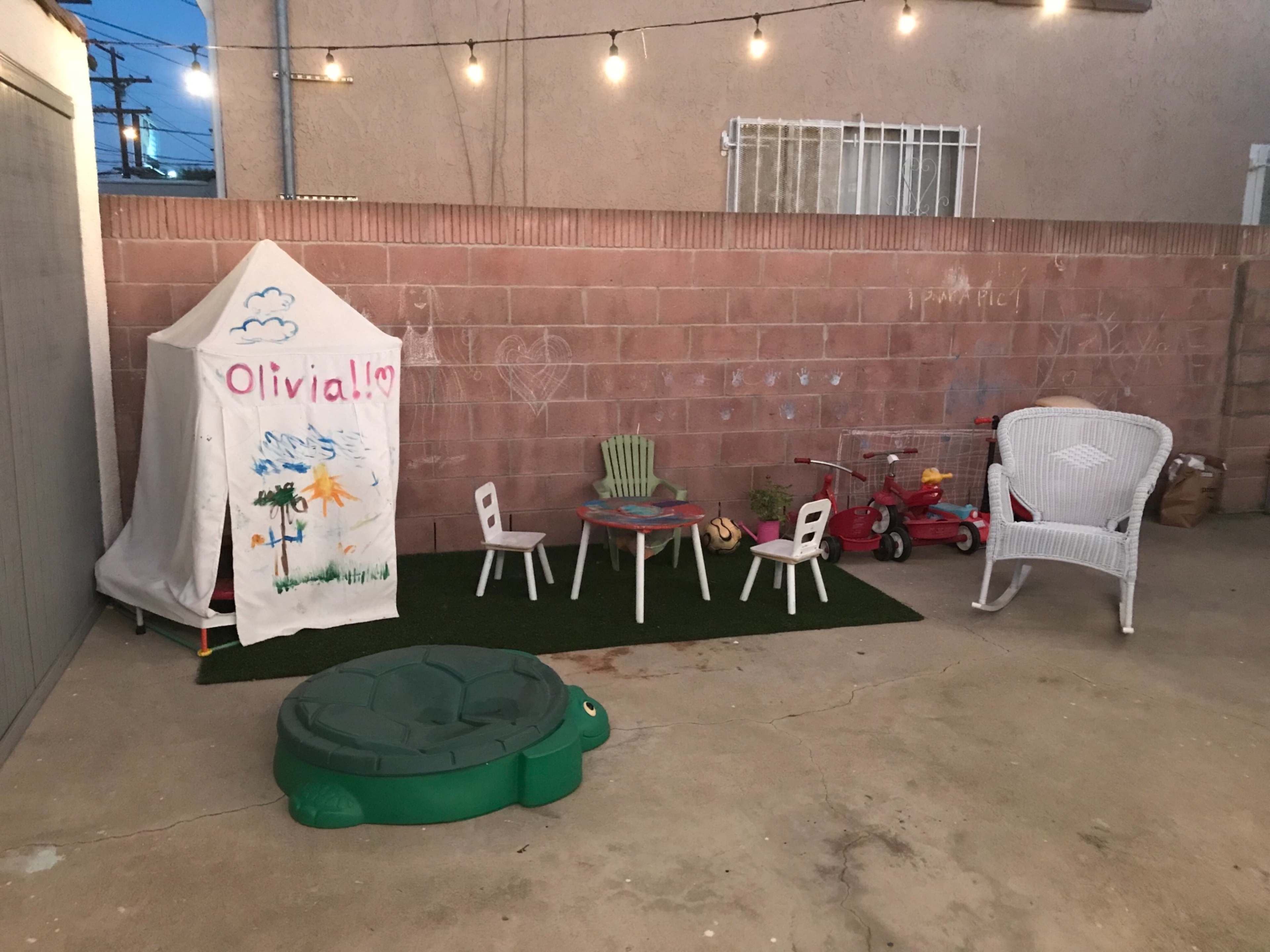 The image shows a small outdoor play area featuring a tent labeled "Olivia!!", a green turtle-shaped sandbox, a small table with chairs, and a tricycle, all set against a wall with a concrete floor.