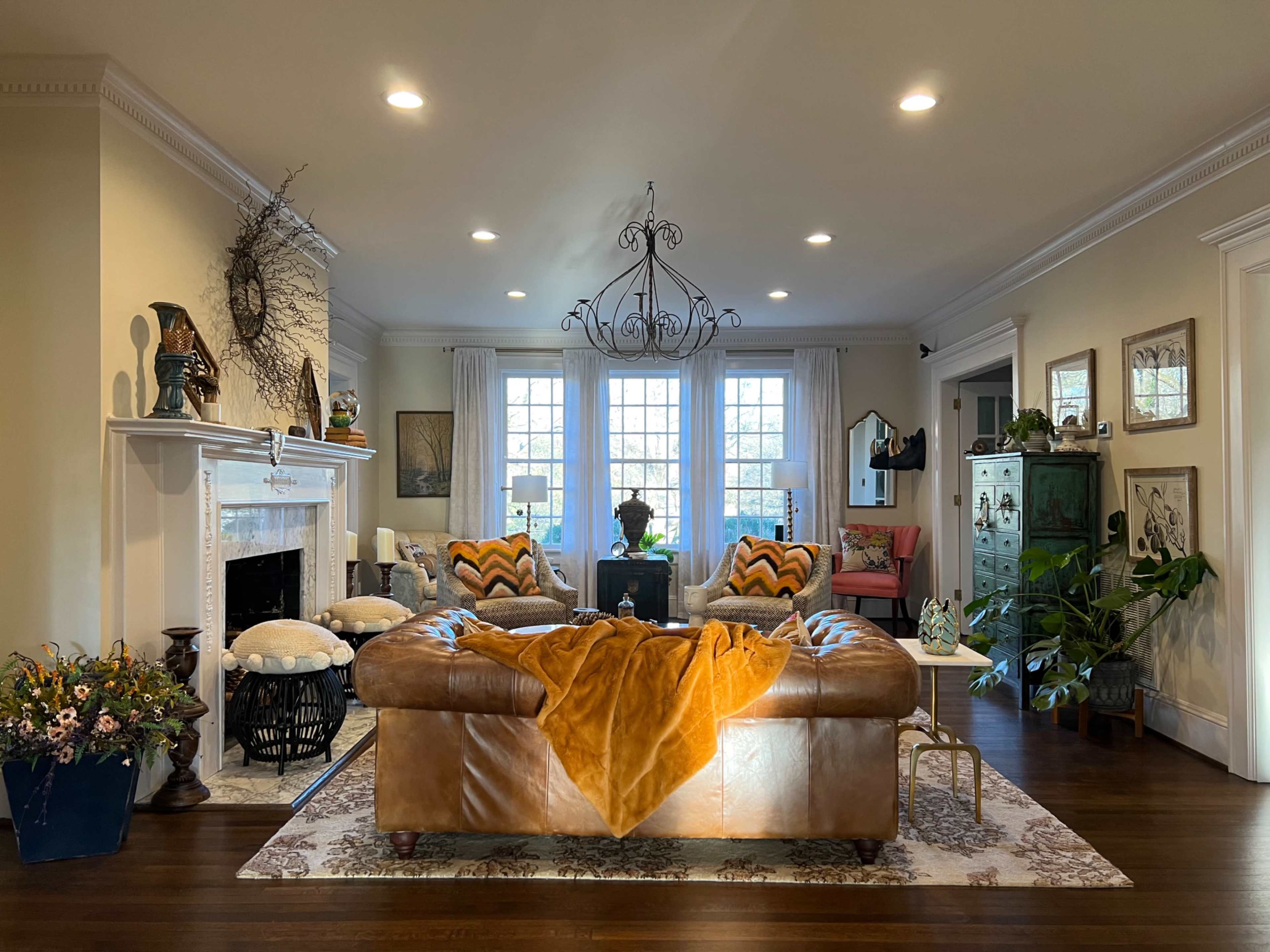 A spacious living room features a brown leather sofa, a decorative chandelier, and large windows letting in natural light, surrounded by various plants and artwork.