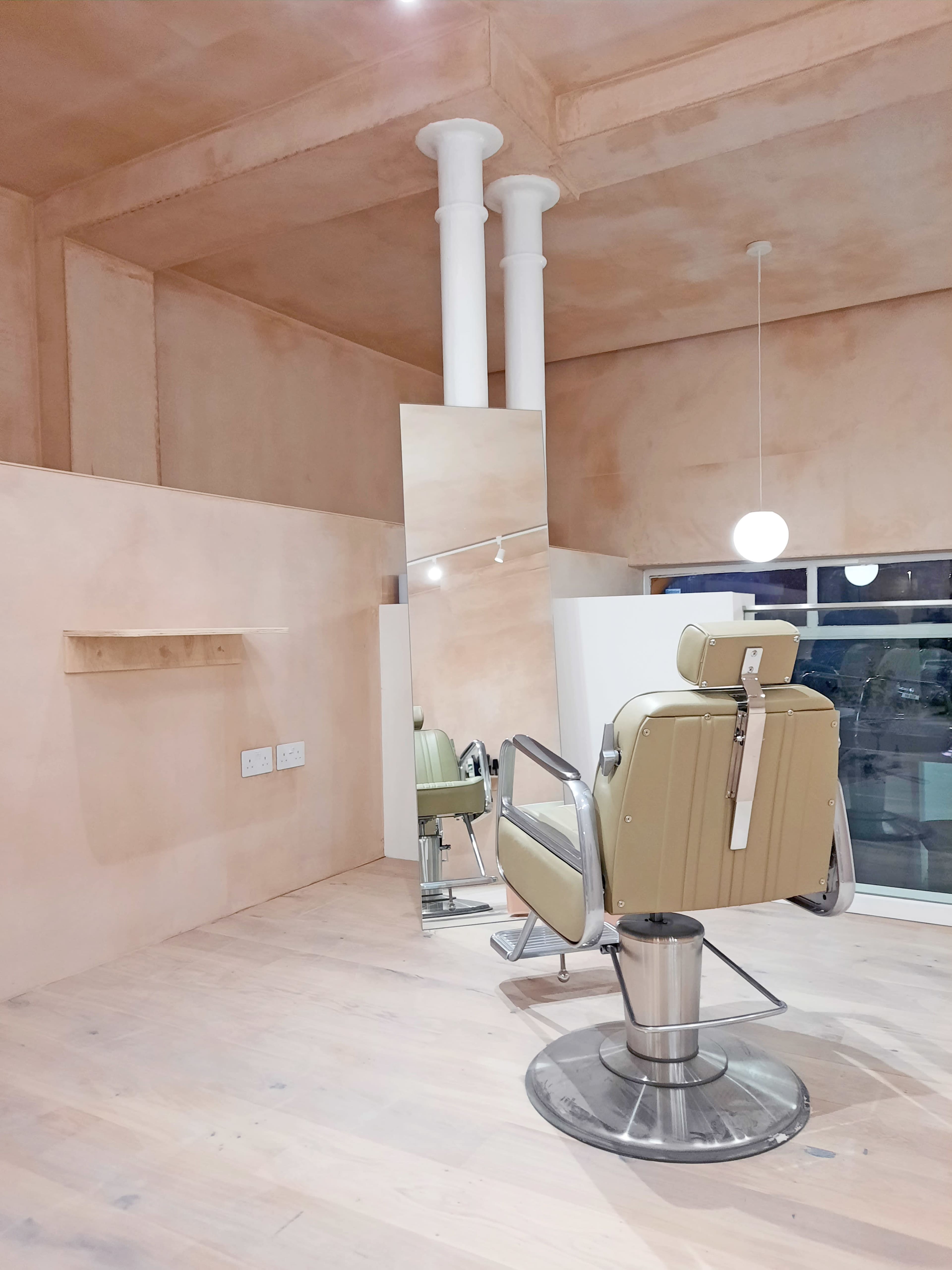 The image shows a minimalist salon interior featuring a single styling chair, a large mirror, and light-colored wooden flooring.