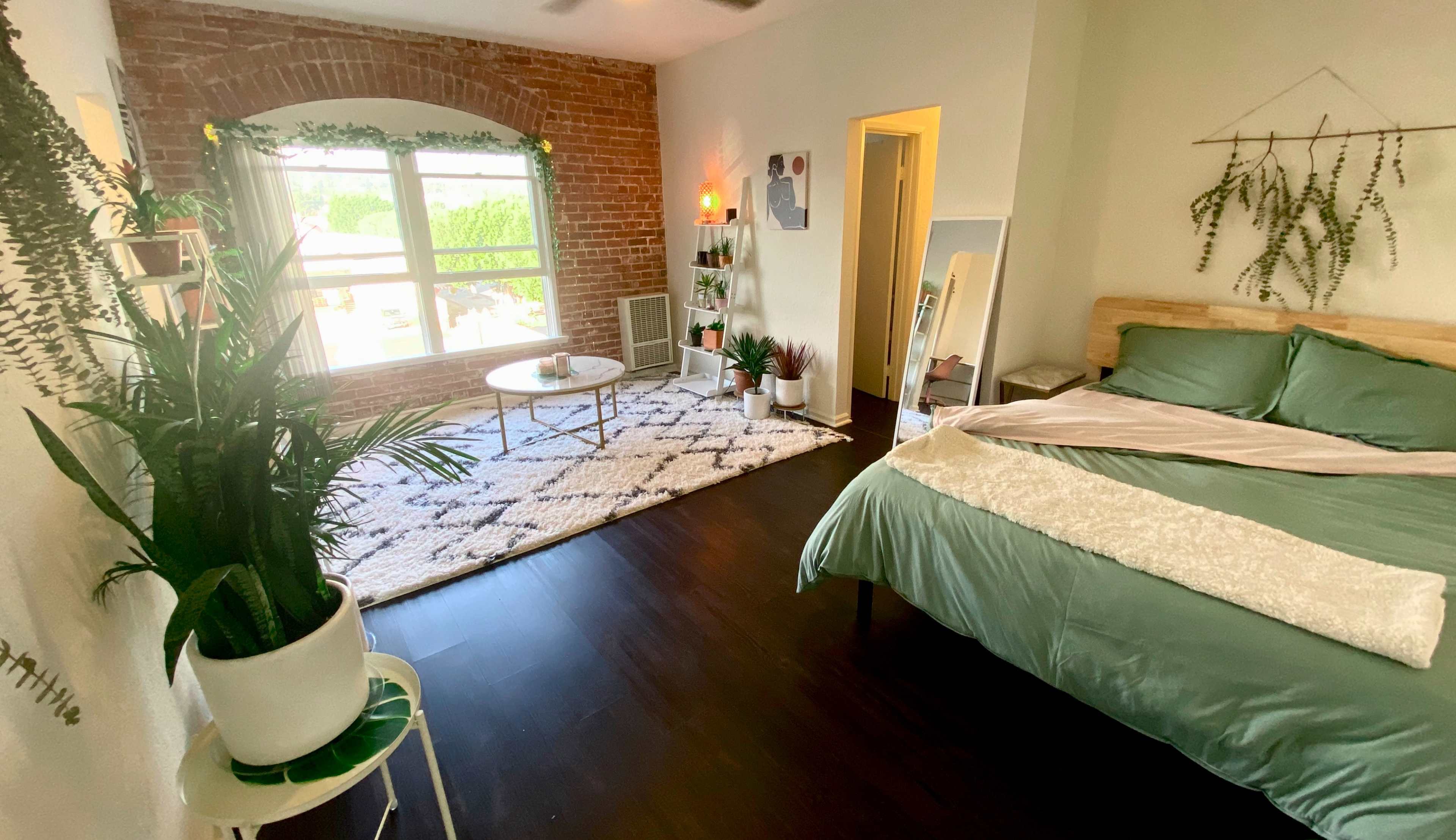 A bedroom with a large window, a bed with green bedding, a seating area with a coffee table, and plants placed around the room.