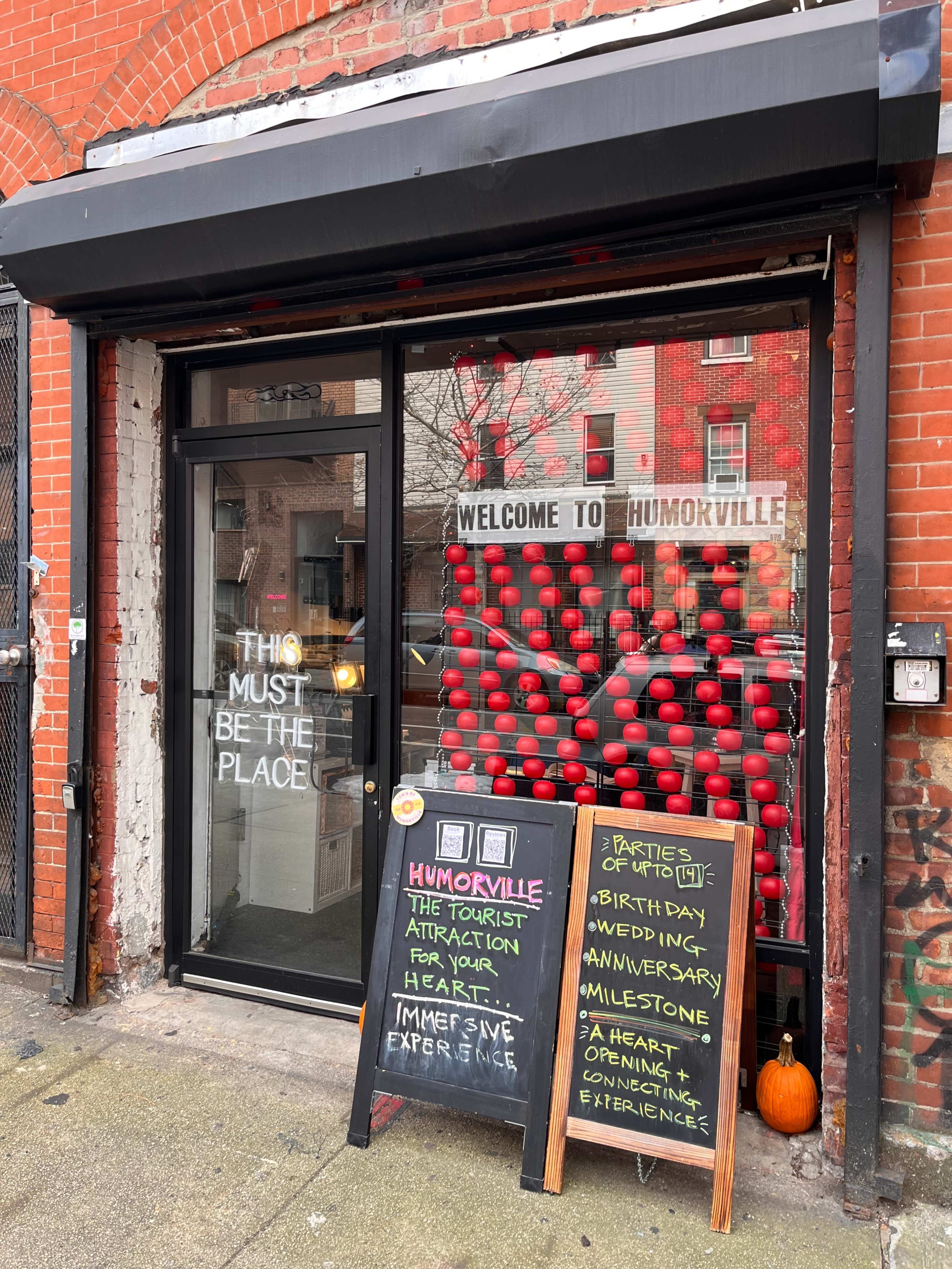 A storefront with large windows displaying red decorations and a sign that reads "Welcome to Humorville," alongside a chalkboard listing event options and a pumpkin at the entrance.