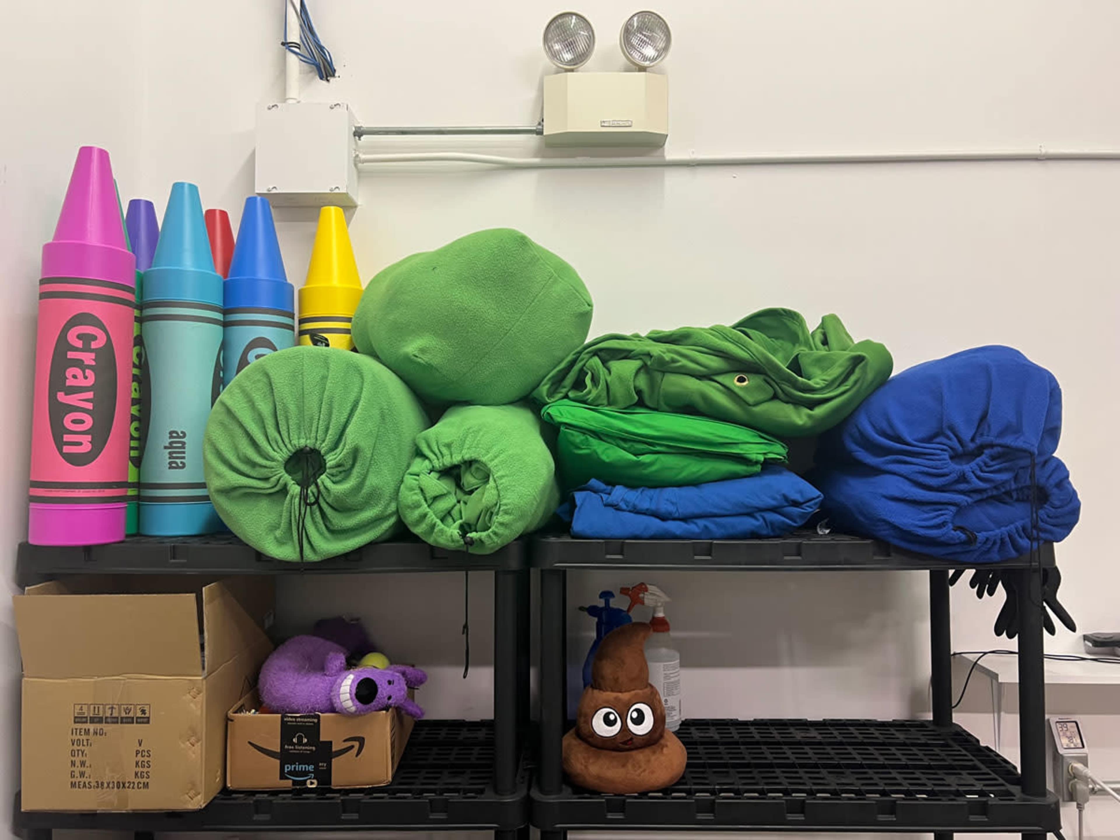 A shelf stacked with colorful plush crayons, green and blue fabric rolls, a toy purple creature, and a brown emoji figure resembling poop.