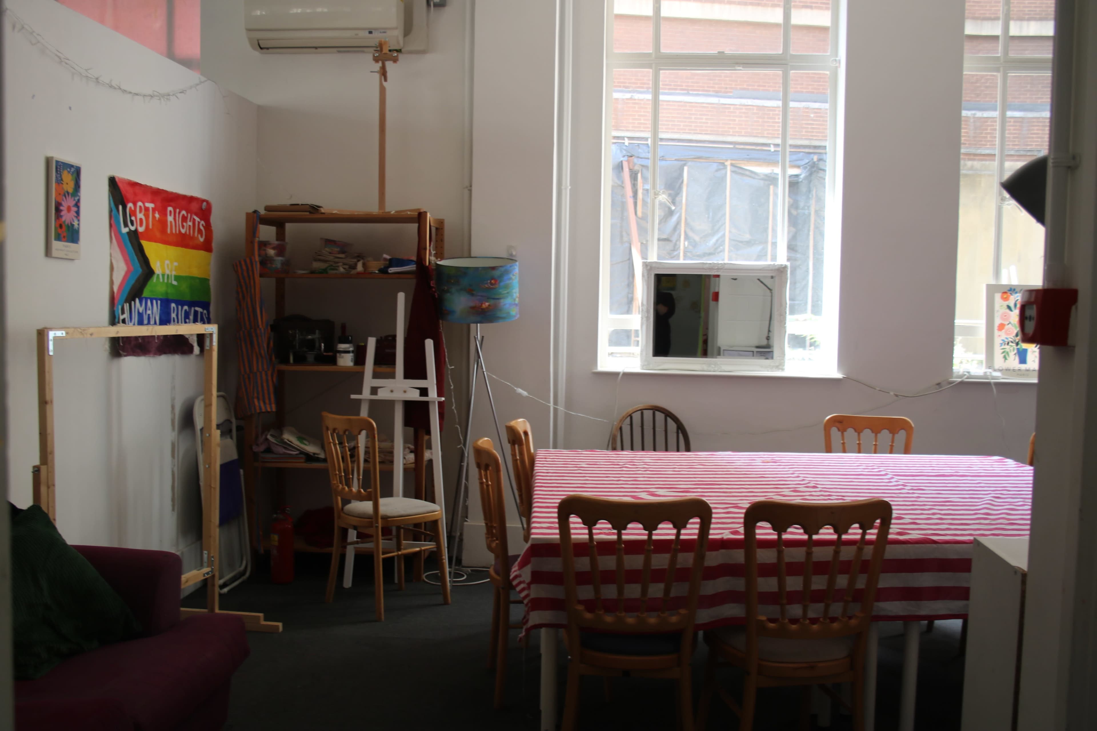 The image shows a room with a large table surrounded by wooden chairs, a purple couch, and various items including an easel, a shelf with books, and a lamp.